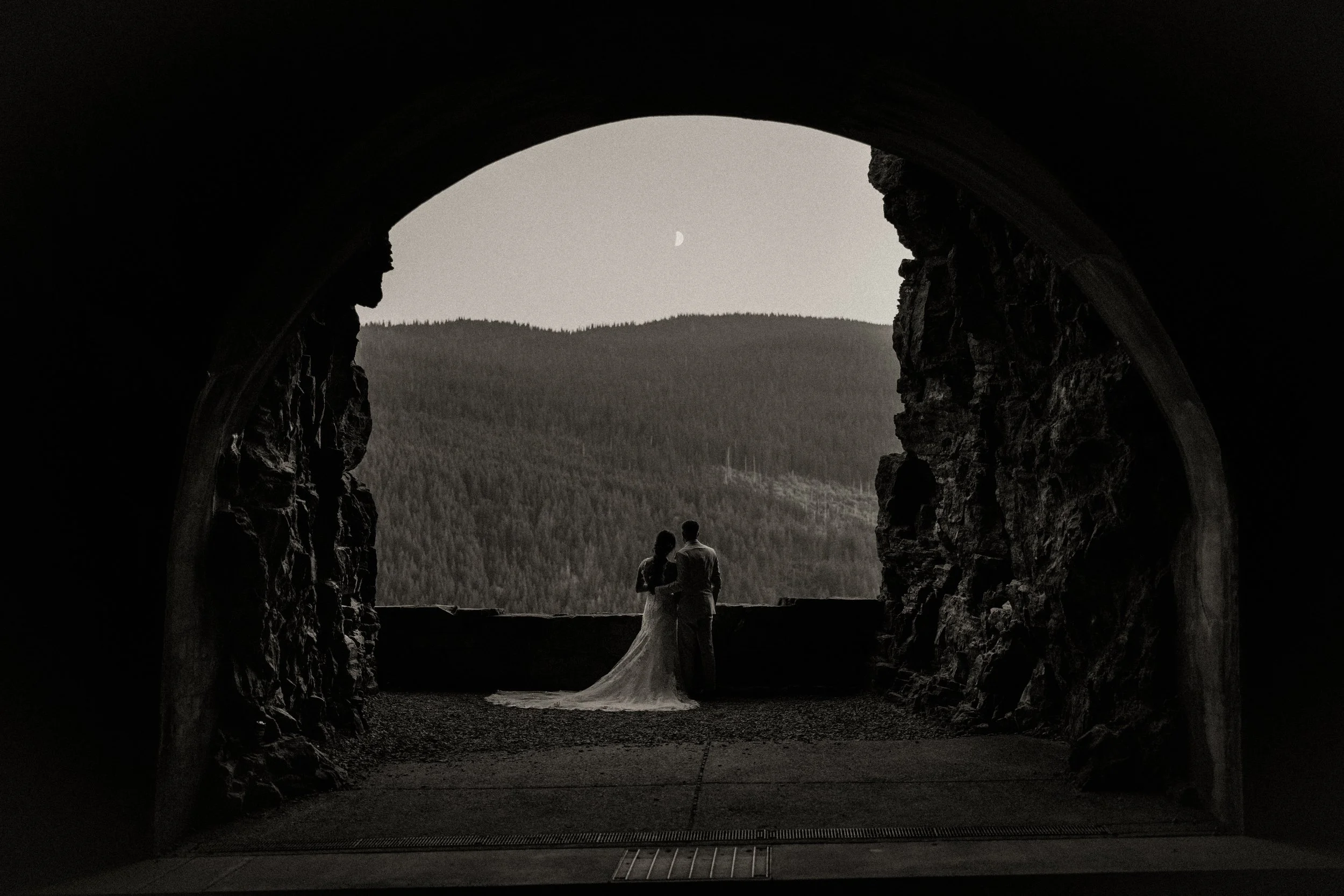 A black and white photo of a couple standing in a cave opening, overlooking a mountain landscape with a visible moon in the sky.
