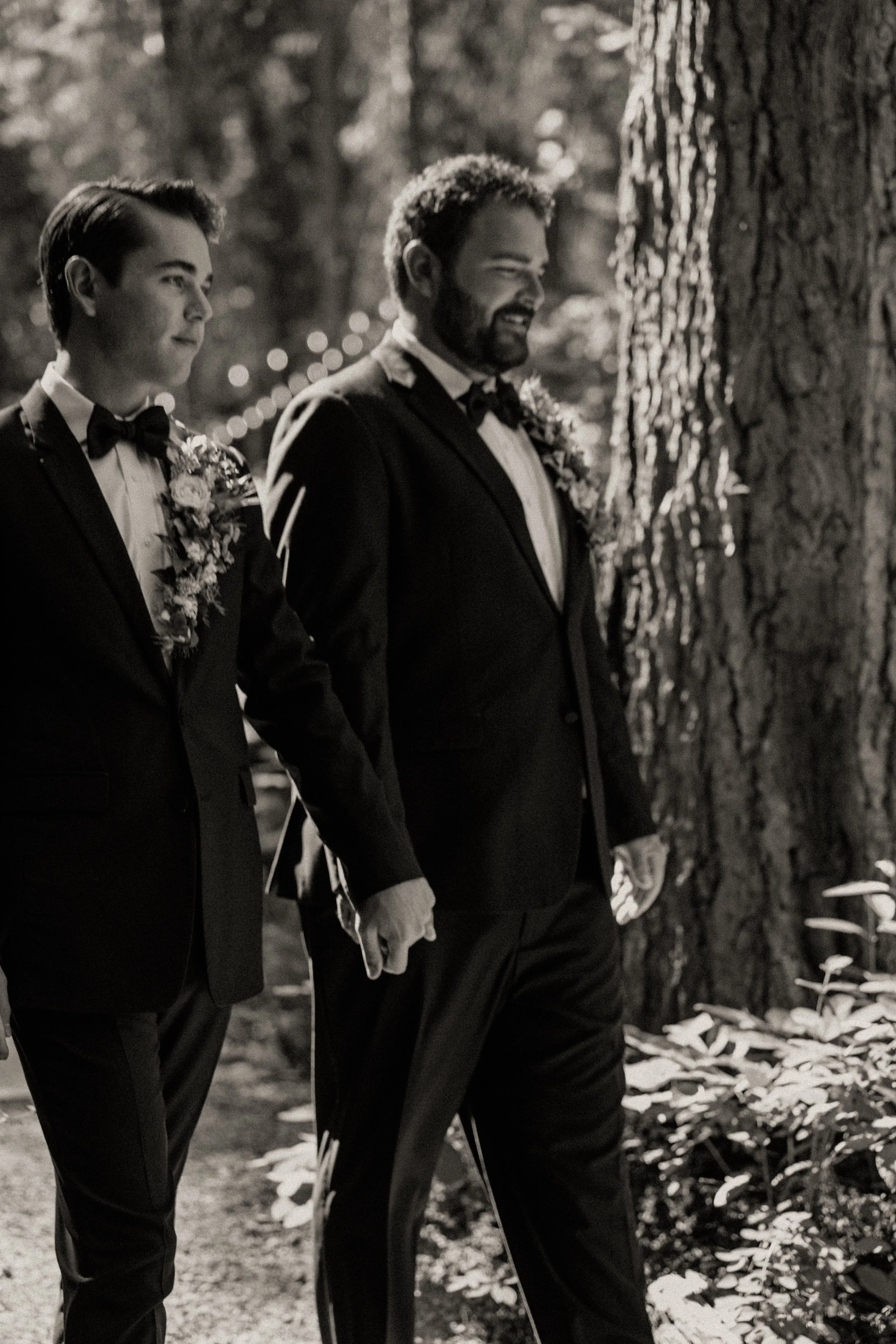 Two men in tuxedos holding hands in a wooded outdoor setting during a wedding.