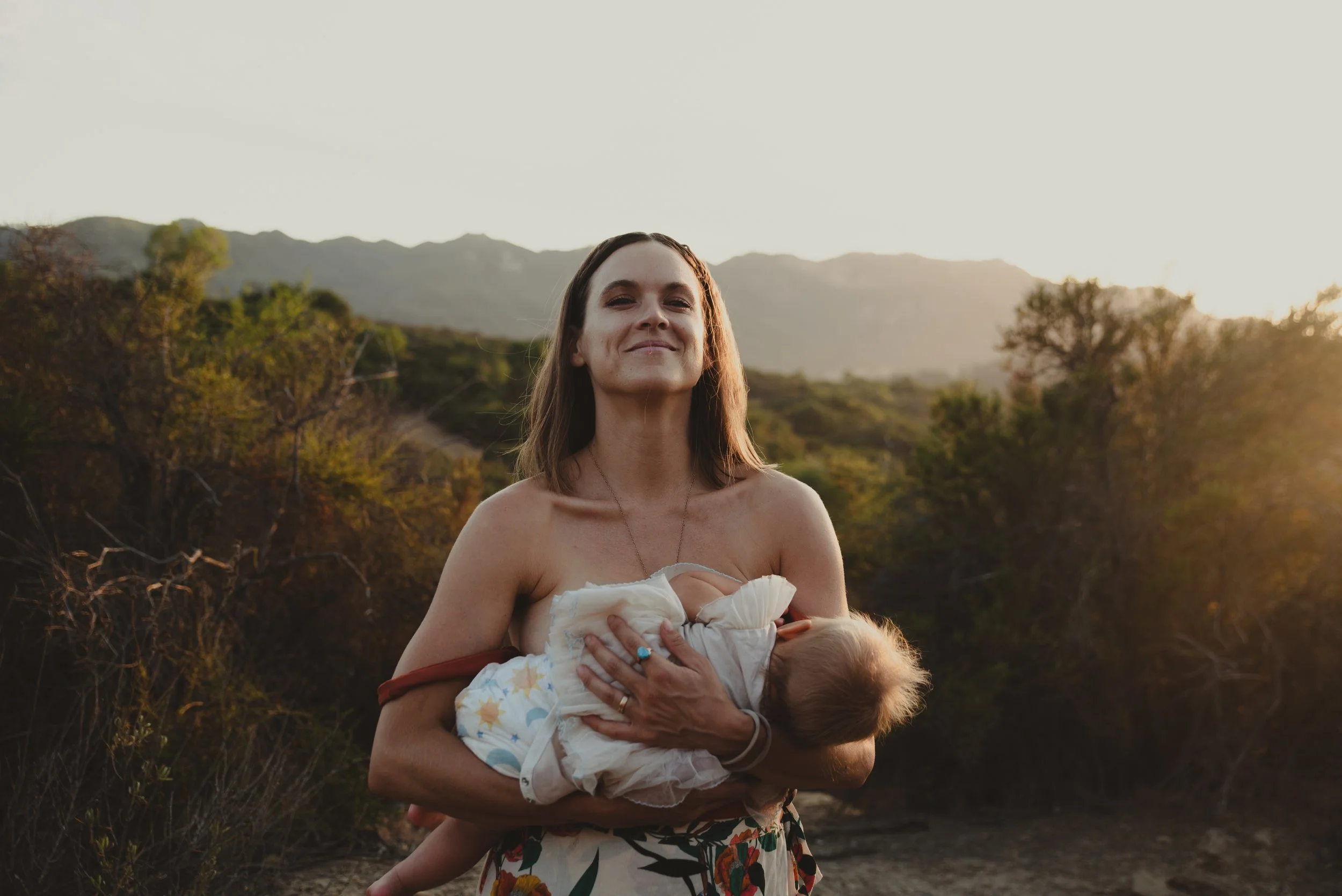 A woman holding a baby outdoors at sunset, with mountains and trees in the background.