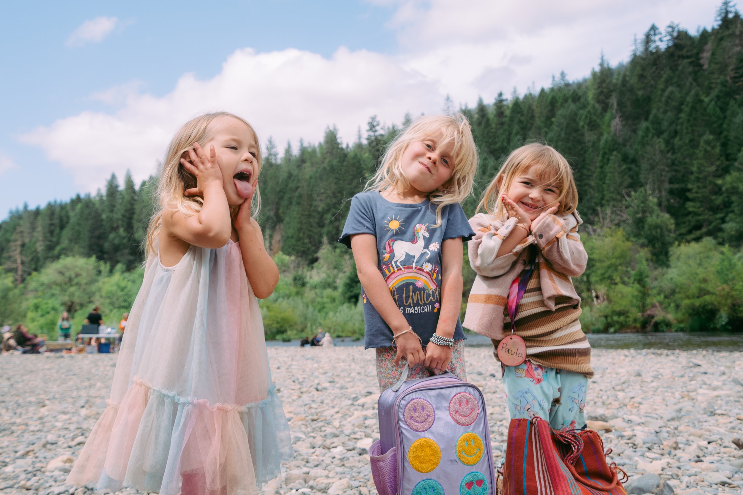 Three young girls standing on a rocky riverbank, making silly faces and smiling, with trees and a river in the background.