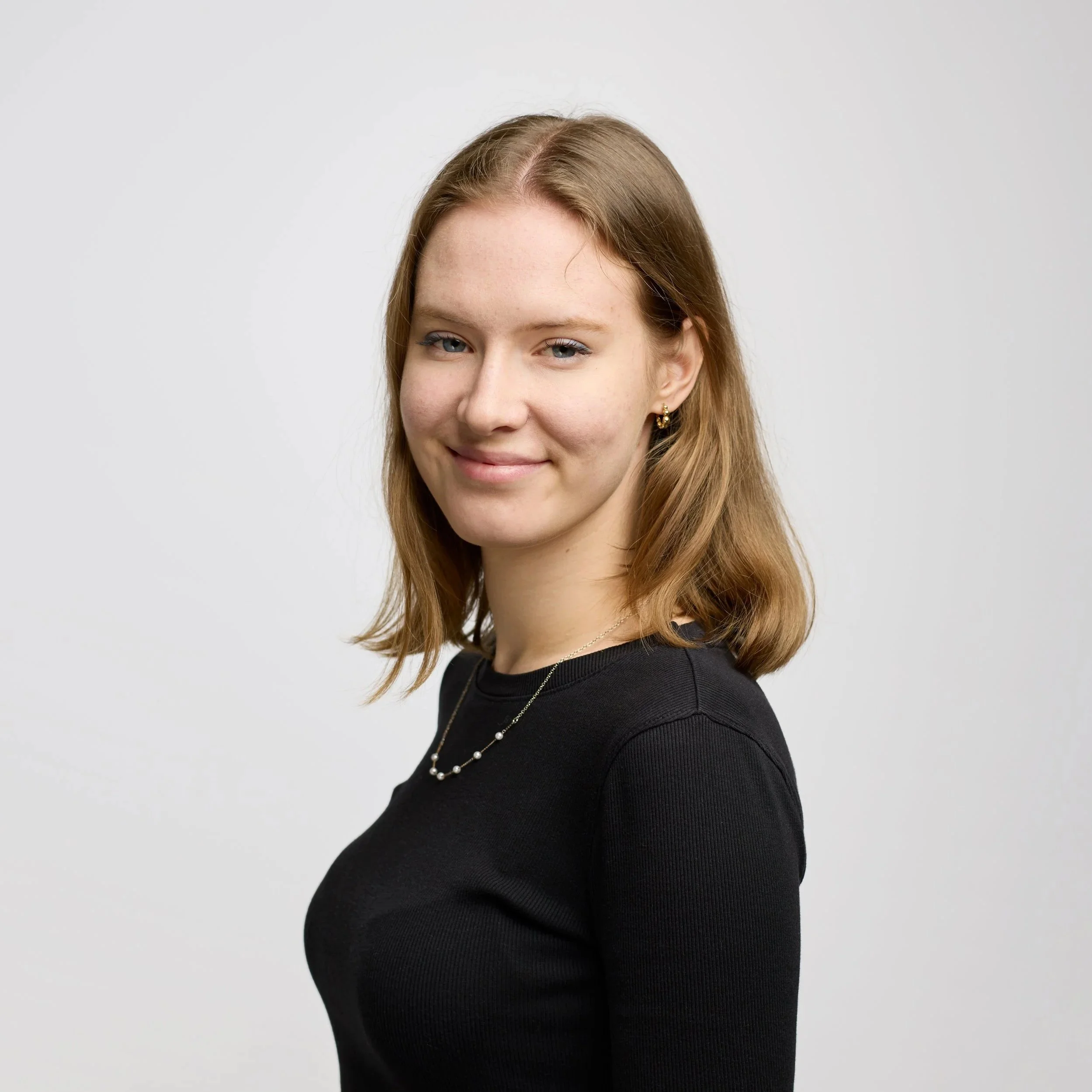 A smiling young woman with shoulder-length blonde hair, wearing a black shirt, earrings, and a gold necklace, against a black background.