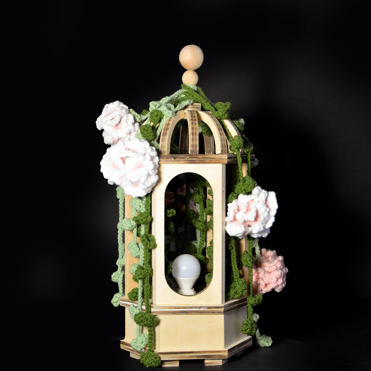 A decorative lantern with artificial pink and white flowers and green crocheted vines surrounding it, displayed against a black background. The lantern has a wooden structure with a lightbulb inside.
