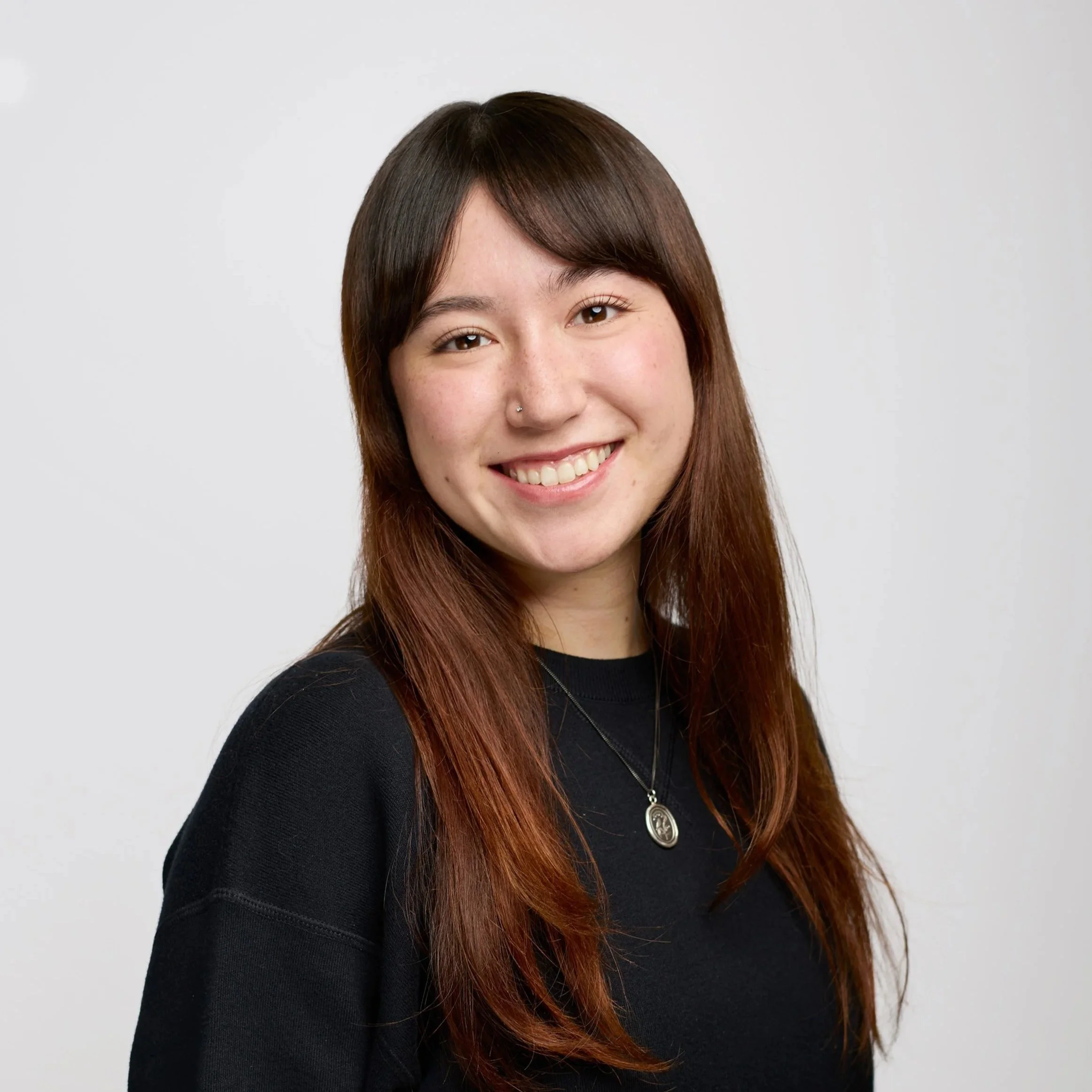 Close-up of a woman with brown hair, wearing a black shirt and a pendant necklace, smiling against a plain white background.