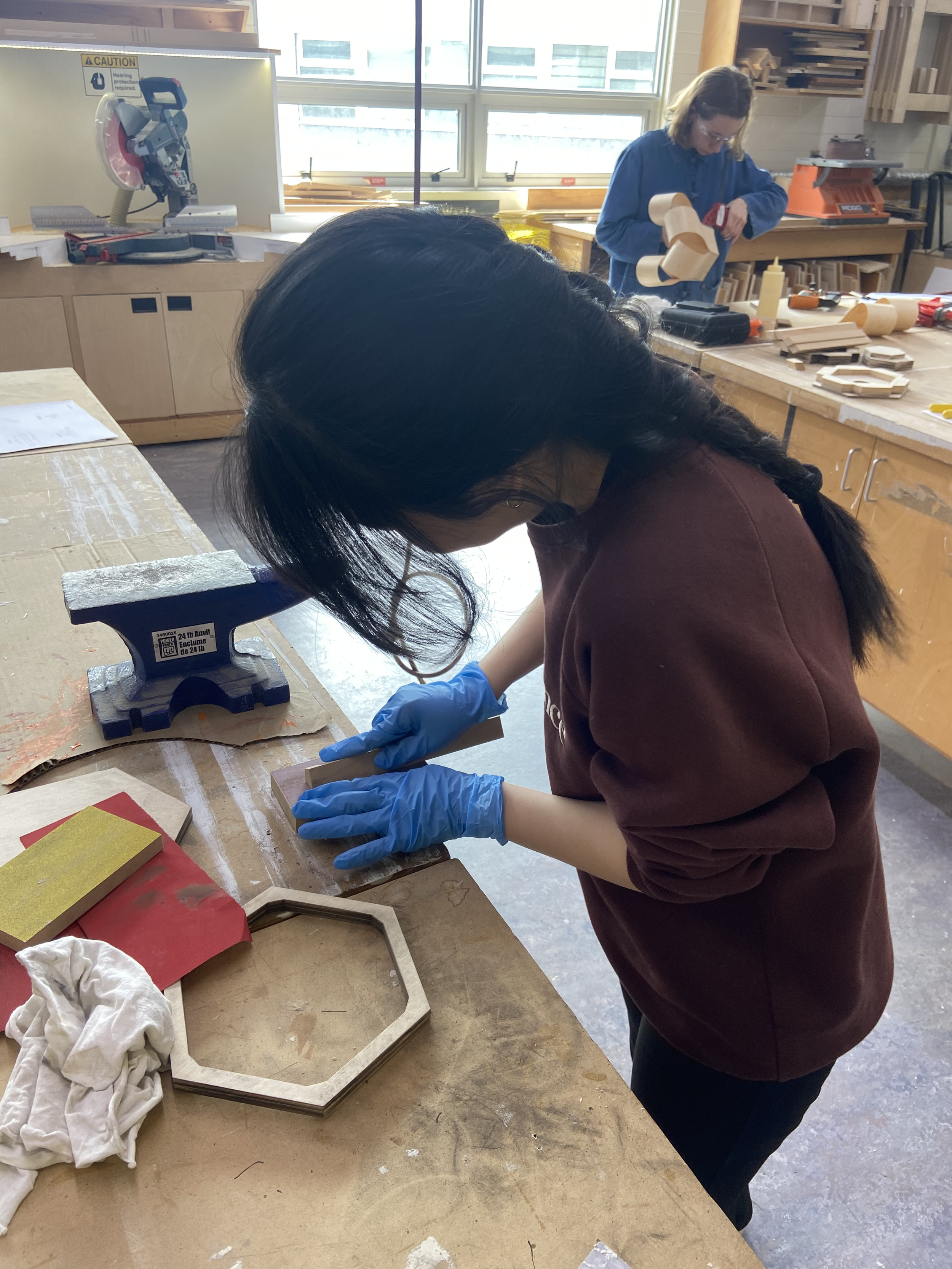 Two women working on woodworking projects in a workshop. One woman with dark hair, wearing a brown shirt and blue gloves, is working on a piece of wood. The other woman with glasses, ginger hair, wearing a blue jacket, is handling multiple wooden pieces. The workspace has various tools, wood pieces, and supplies on the tables.
