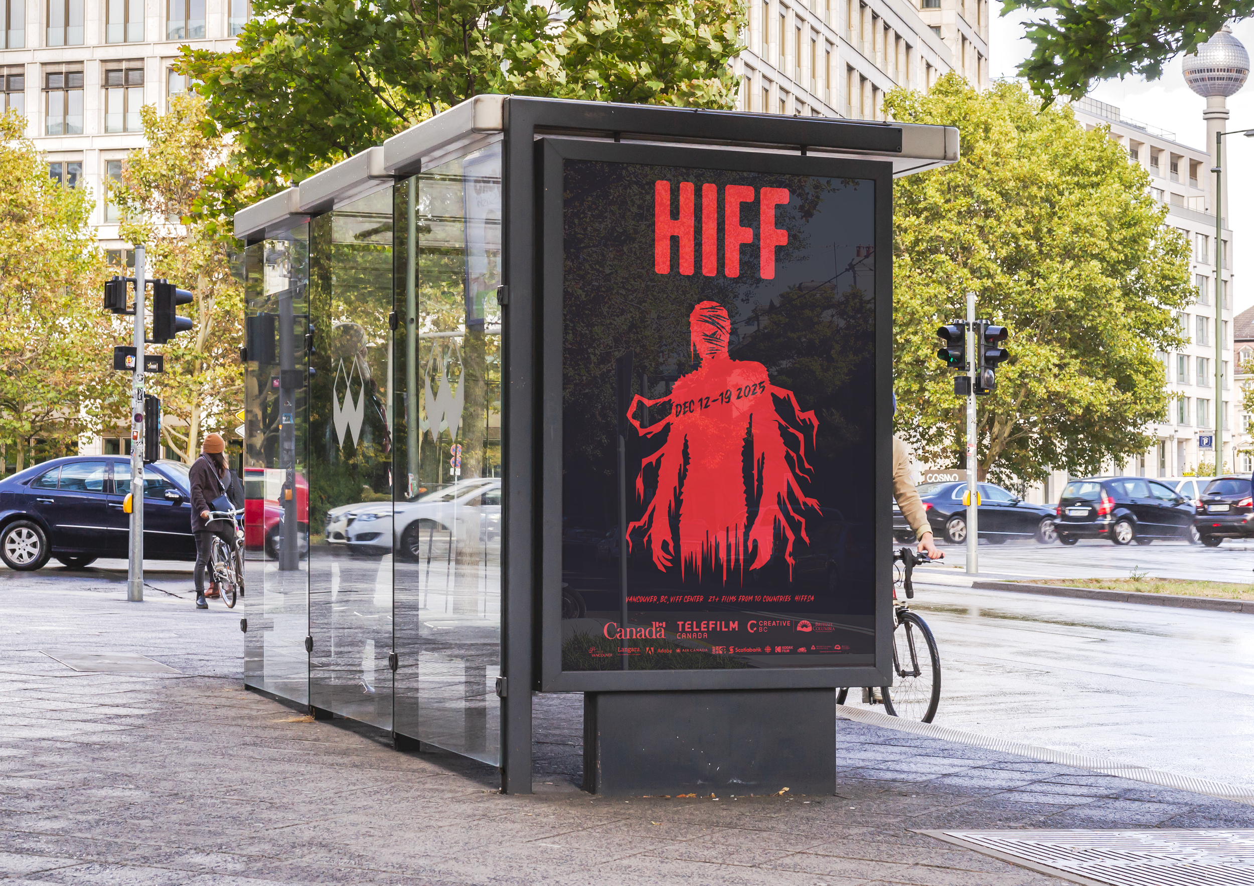 A digital billboard at a bus stop displaying an advertisement for HIFF, Vancouver, BC, with the dates December 12-19, 2025, and logos of sponsors and partners at the bottom. The surrounding area includes trees, buildings, and people with bicycles.