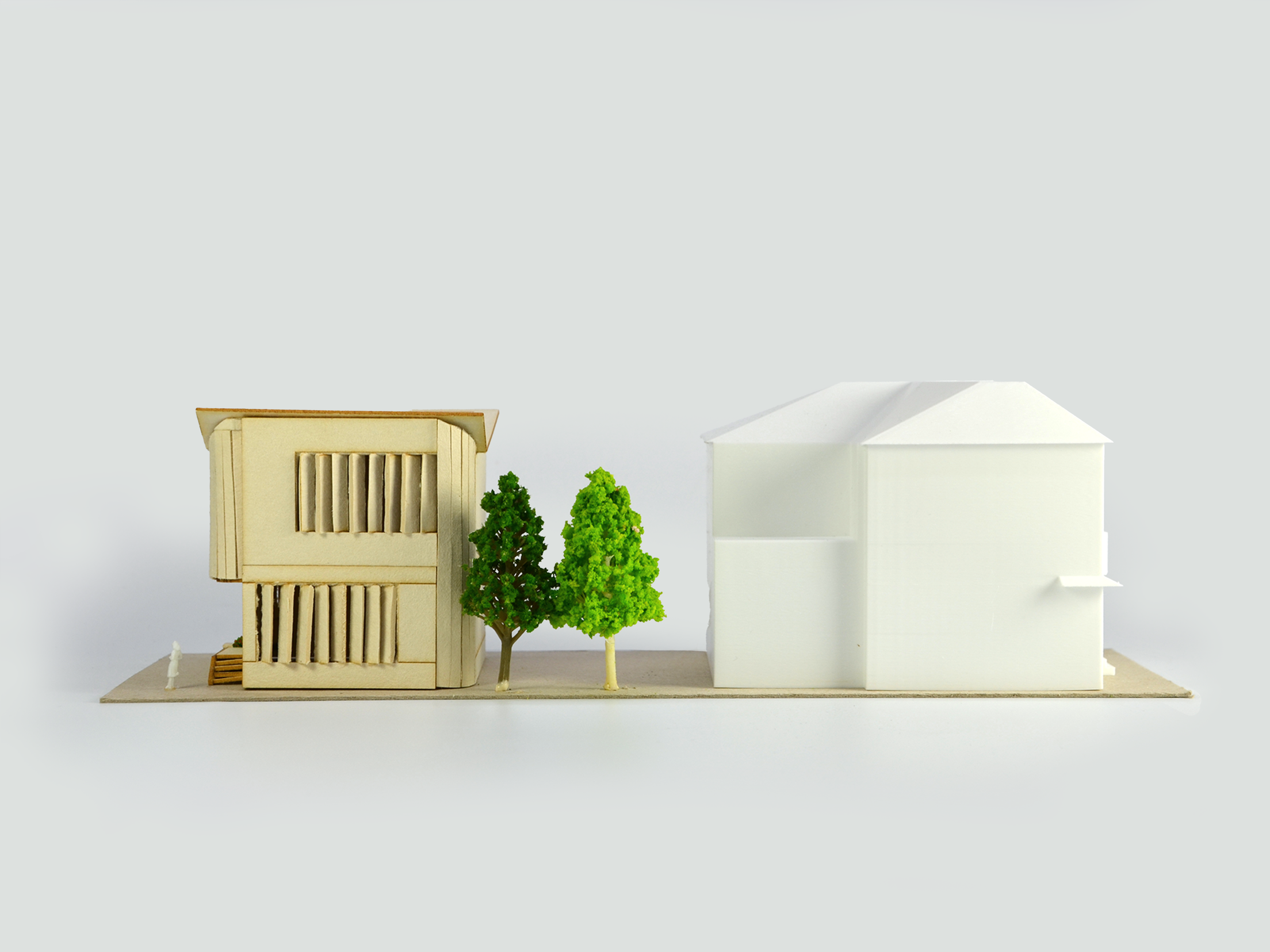 Architectural model featuring two modern houses with trees in front, the house on the left is made of wood with vertical slats and a small staircase, and the house on the right is white with a gabled roof, all set on a flat base