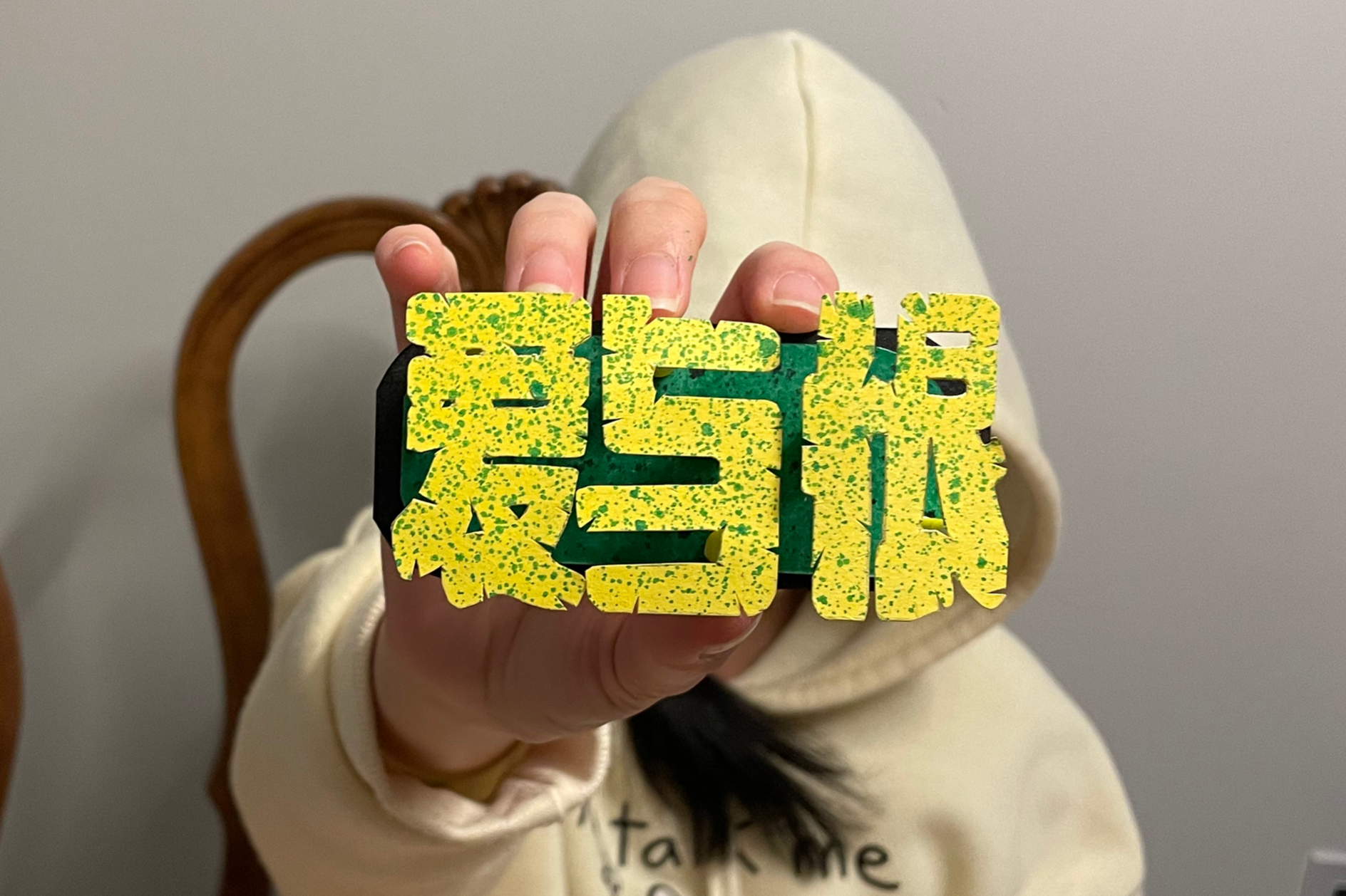 Person holding a yellow and black sign with the word "SISTER" in bold letters. The person is wearing a cream-colored hoodie and is sitting in front of a wooden chair with a light gray wall background.