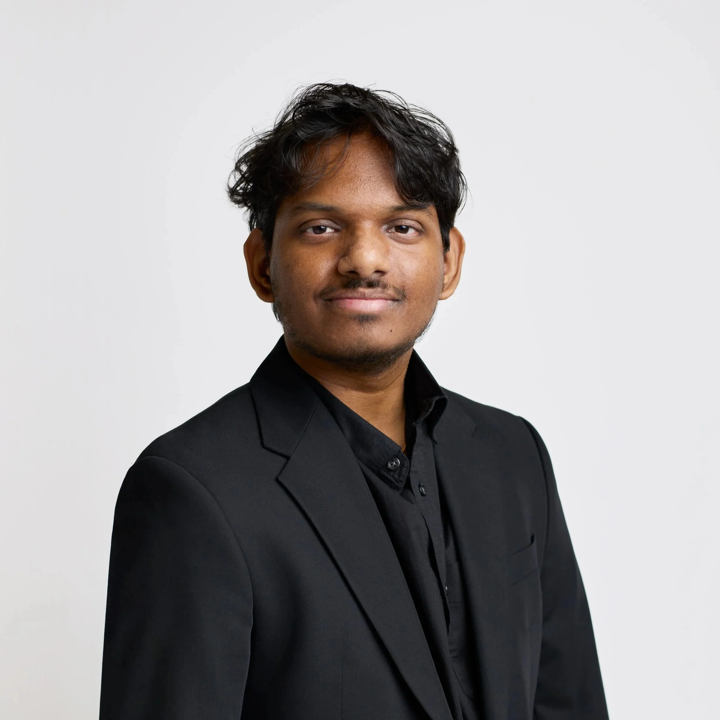 Portrait of a young smiling man, with dark short hair and facial hair, wearing a black suit, on a white background.
