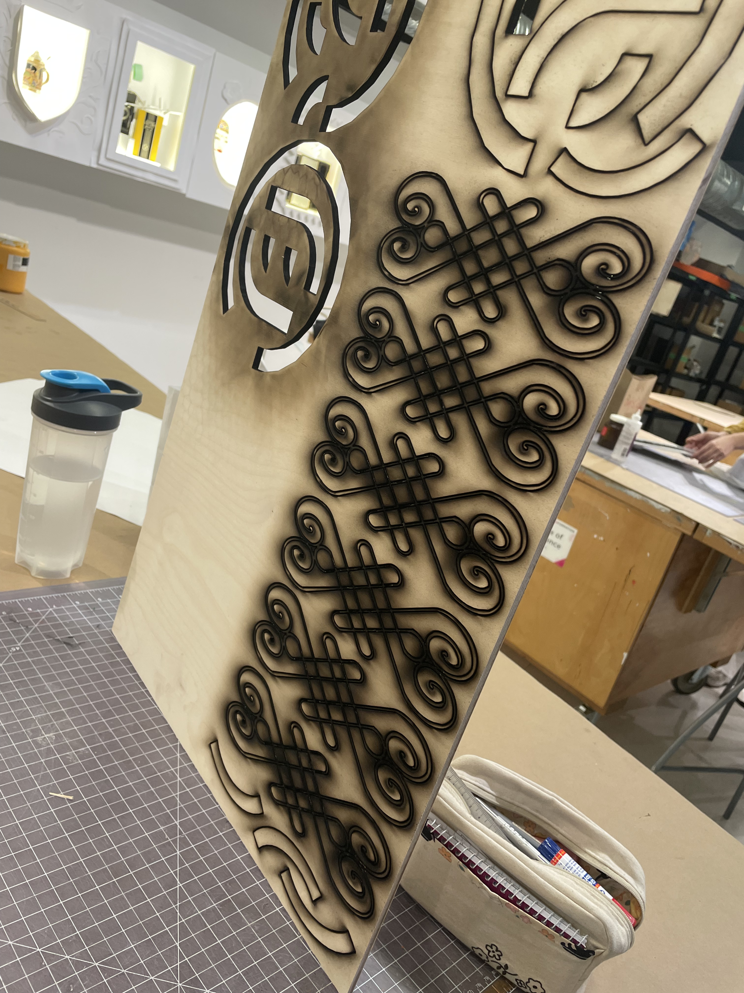 A wooden board with black metal decorative cutouts featuring intricate scrollwork and patterns. The background shows a workspace with various crafting tools, a water bottle, and a white bag.