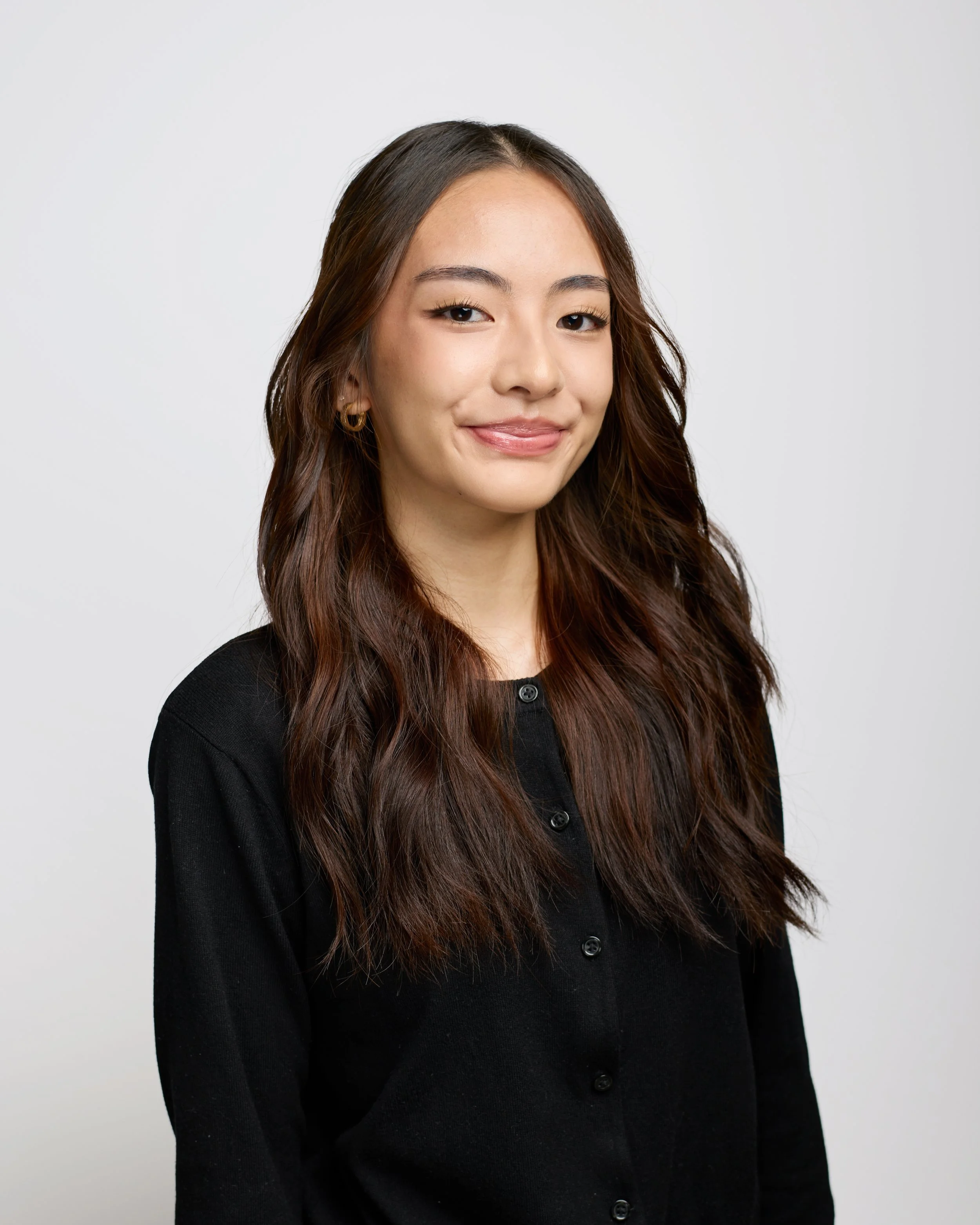 A young woman with long wavy brown hair wearing a black top and gold loop earrings, smiling softly against a plain white background.