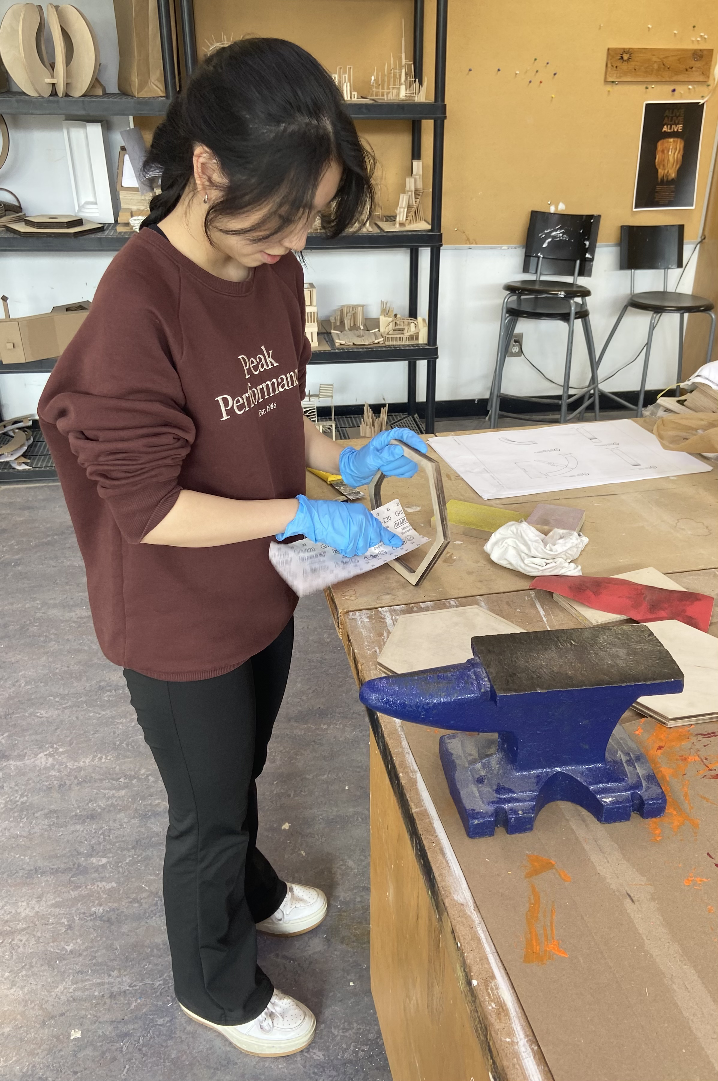 A woman in a maroon sweatshirt and black pants is working in a workshop, wearing blue gloves. She is standing at a wooden workbench, inspecting an object. There are sketches, pieces of wood, and a blue vise on the table. In the background, there are shelves with small wooden structures, a bulletin board, and two black chairs with metal legs against a yellow wall.