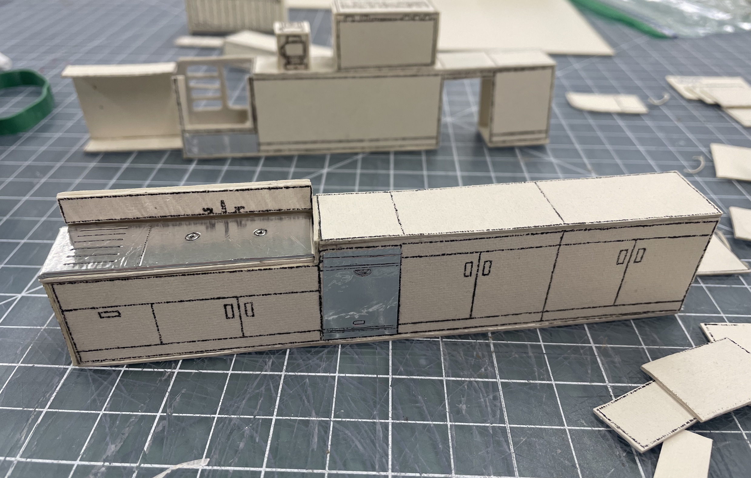 Unassembled miniature furniture pieces made of white cardboard and metal, laid out on a black cutting mat with grid lines. The pieces have black hand-drawn details resembling cabinet doors and drawers.
