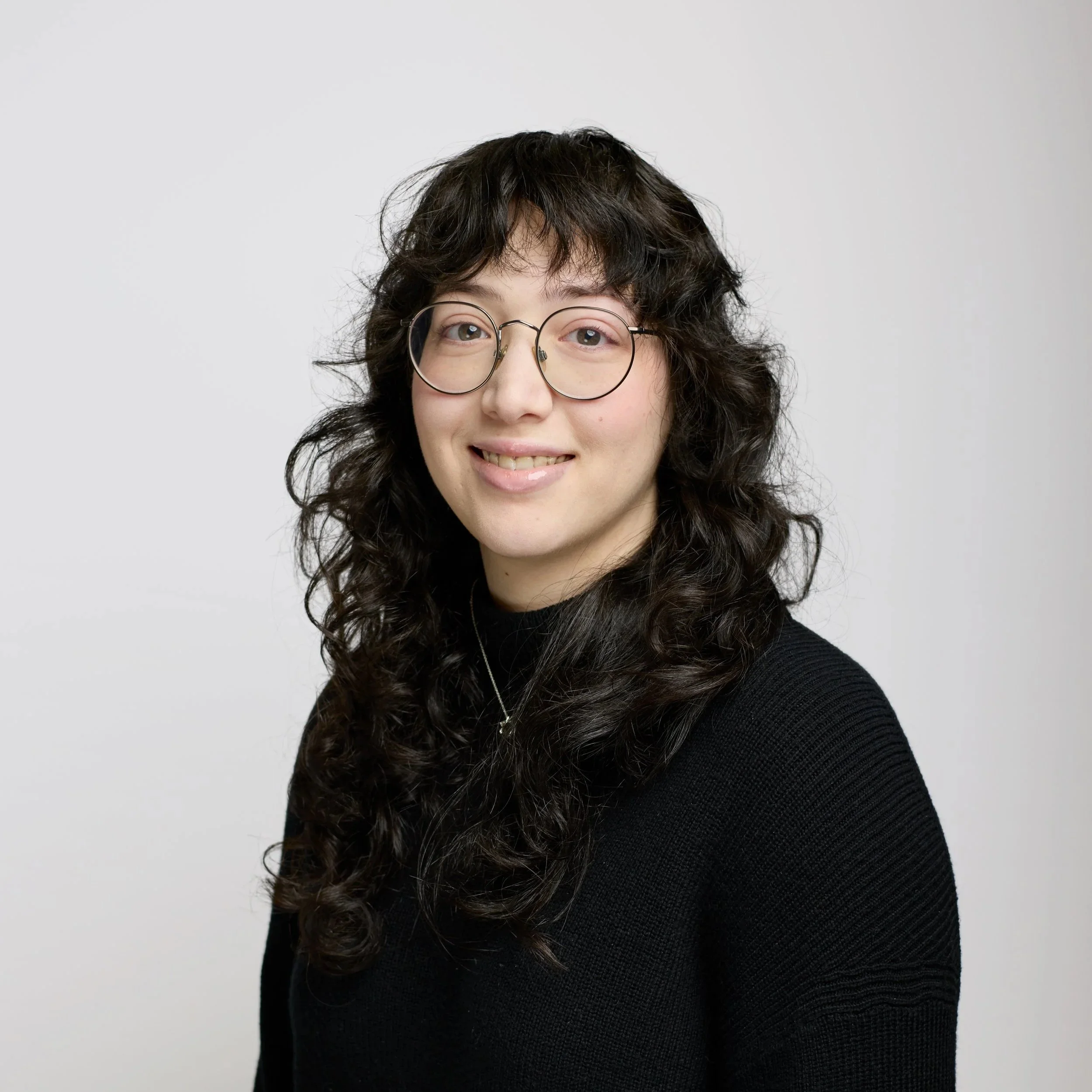 Portrait of a young woman with long blonde hair, smiling, wearing a black top and a silver heart necklace, against a black background.