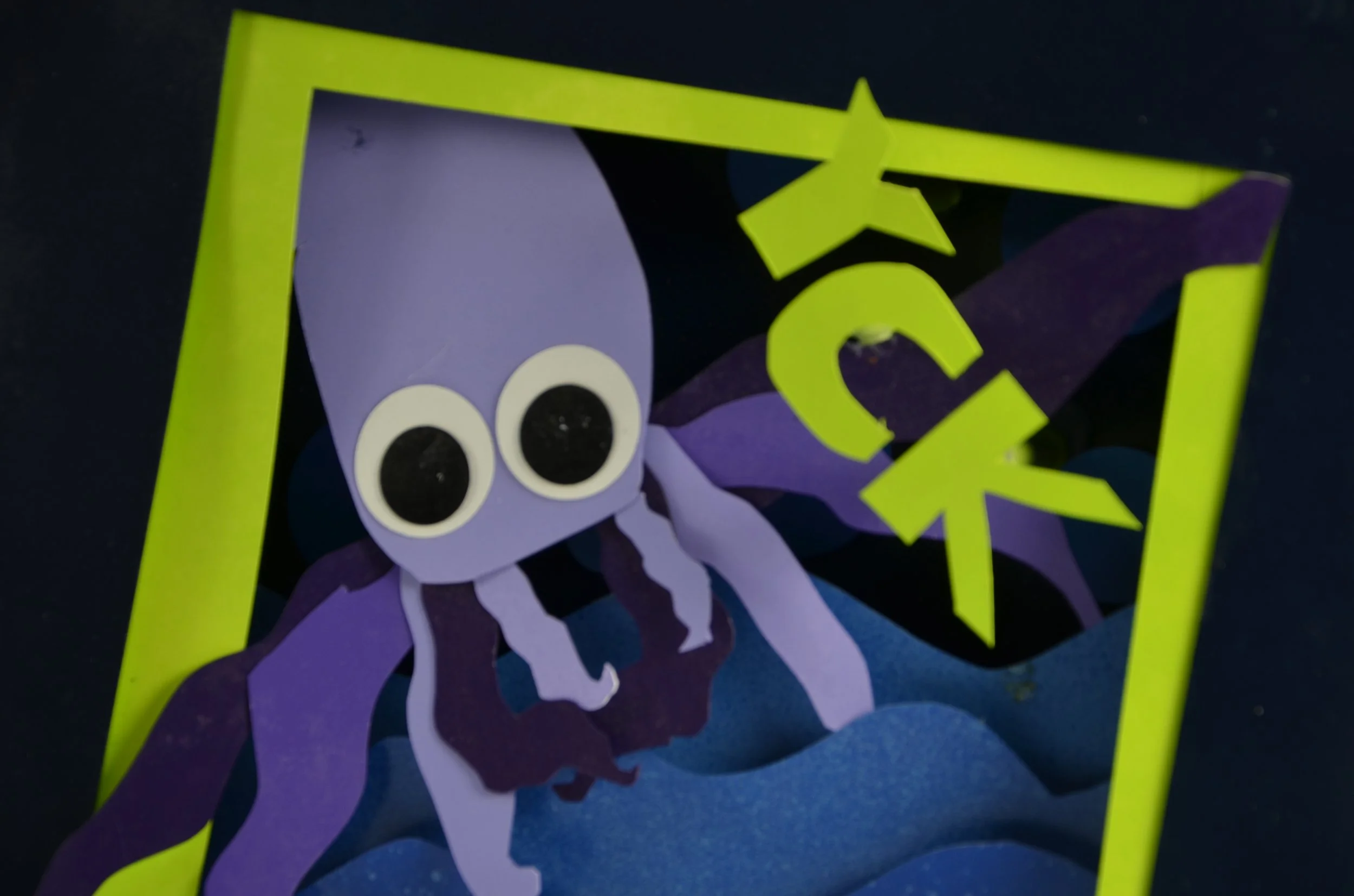 Colorful paper cutouts of a purple octopus with big white eyes and a green star on its head, placed inside a yellow photo frame.