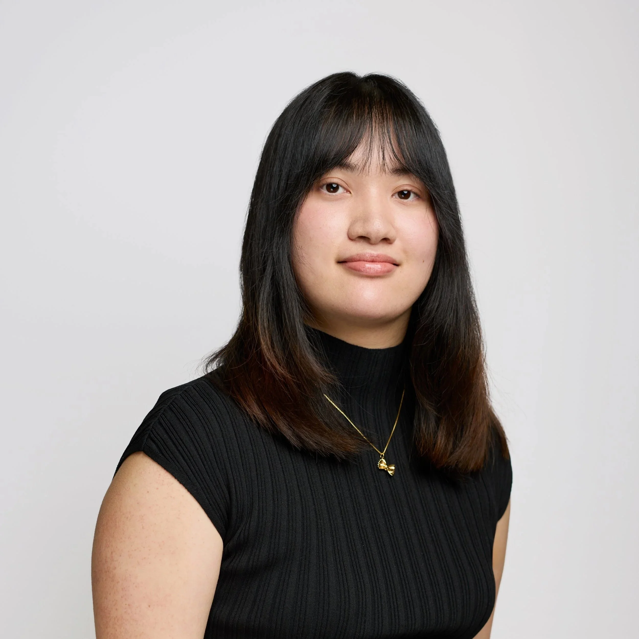 Portrait of a young woman with long auburn hair, wearing a black top and gold necklace, smiling softly against a black background.
