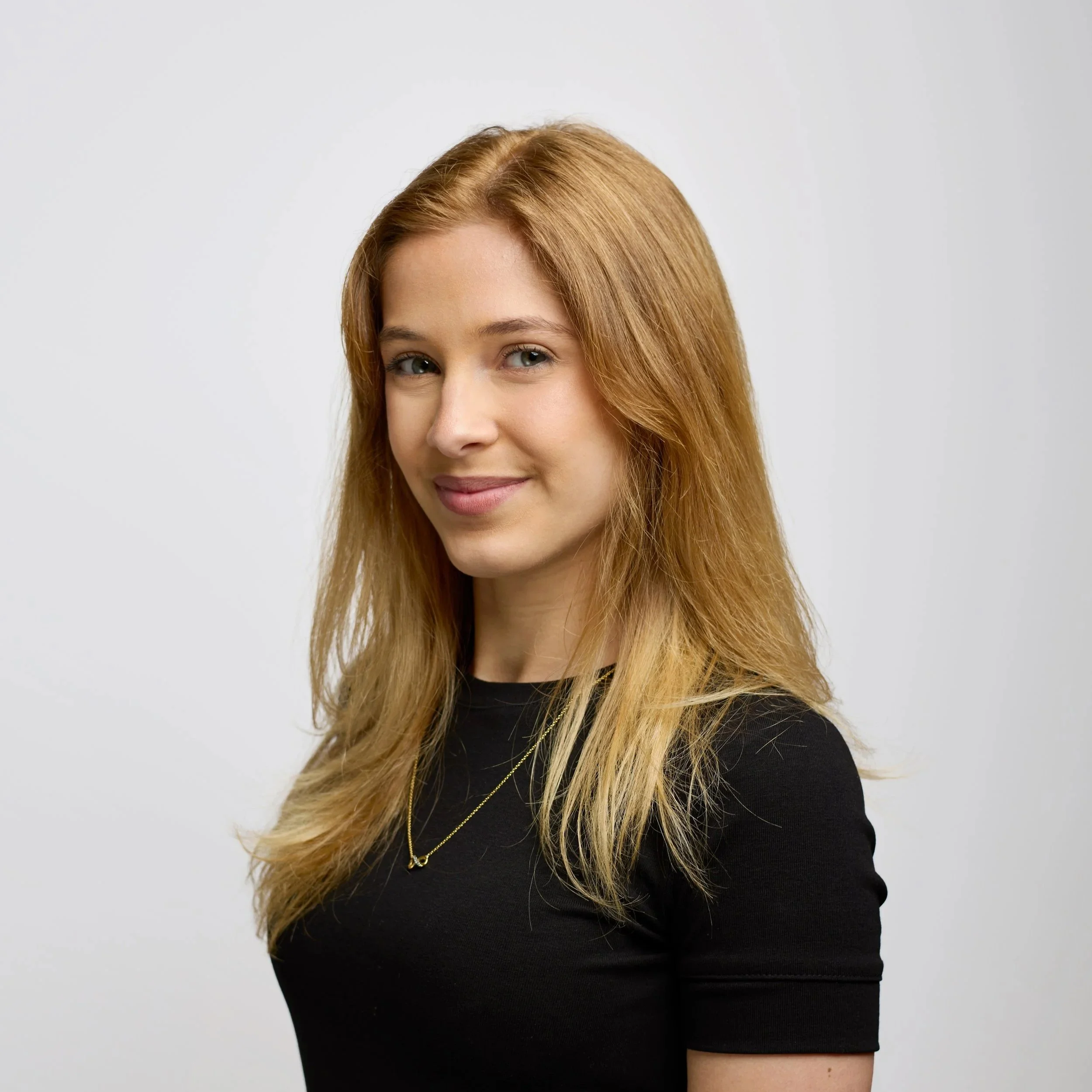 A young woman with medium length blonde hair, smiling at the camera, wearing a black t-shirt and a gold necklace, on a white background.