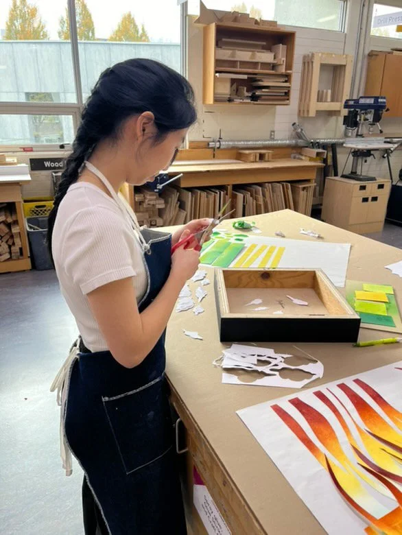 A woman standing at a workbench cutting paper with scissors in a woodworking or craft room. There are colorful paper strips and cutouts on the table, and shelves with wood pieces and tools in the background.