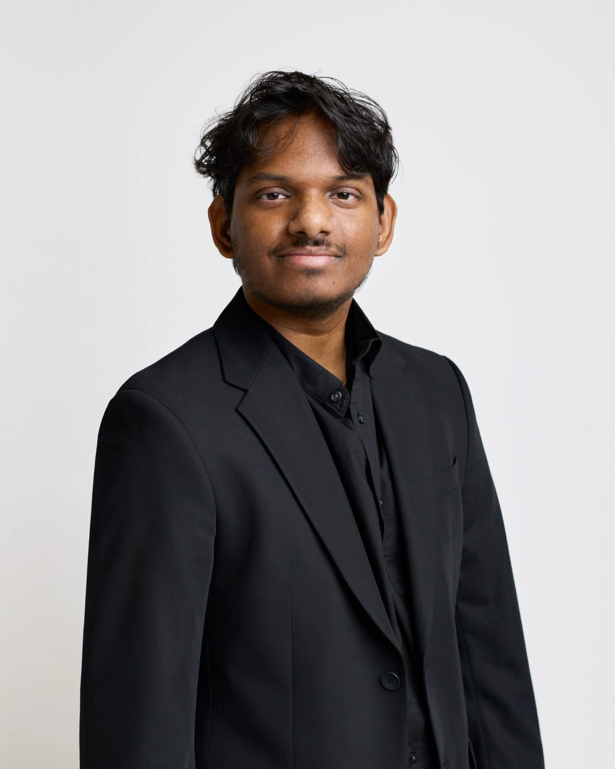 A man in a black suit and black shirt posing against a plain white background.