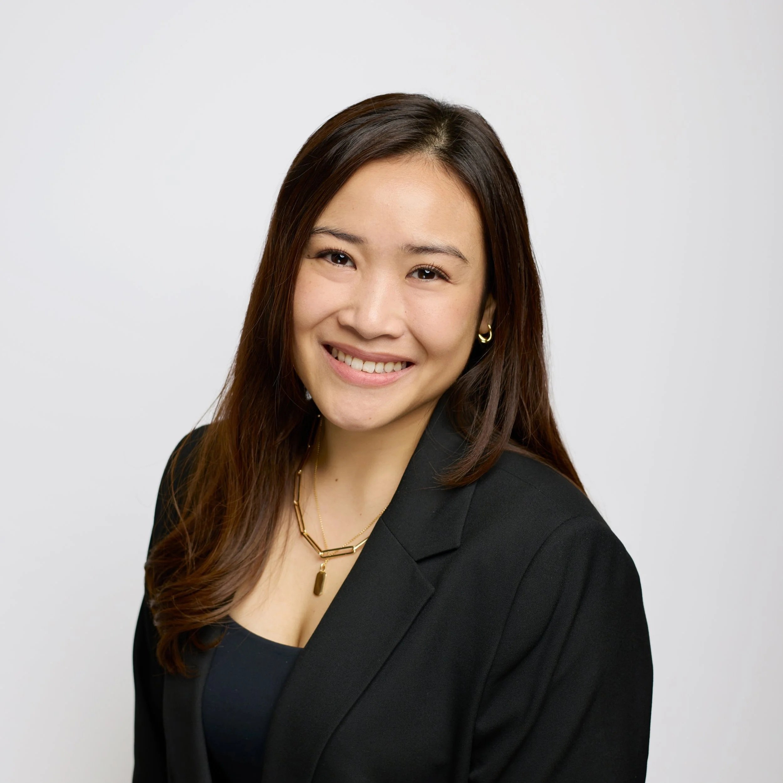 Smiling young woman with long dark hair and bangs, wearing a white top and gold necklace, against a black background.