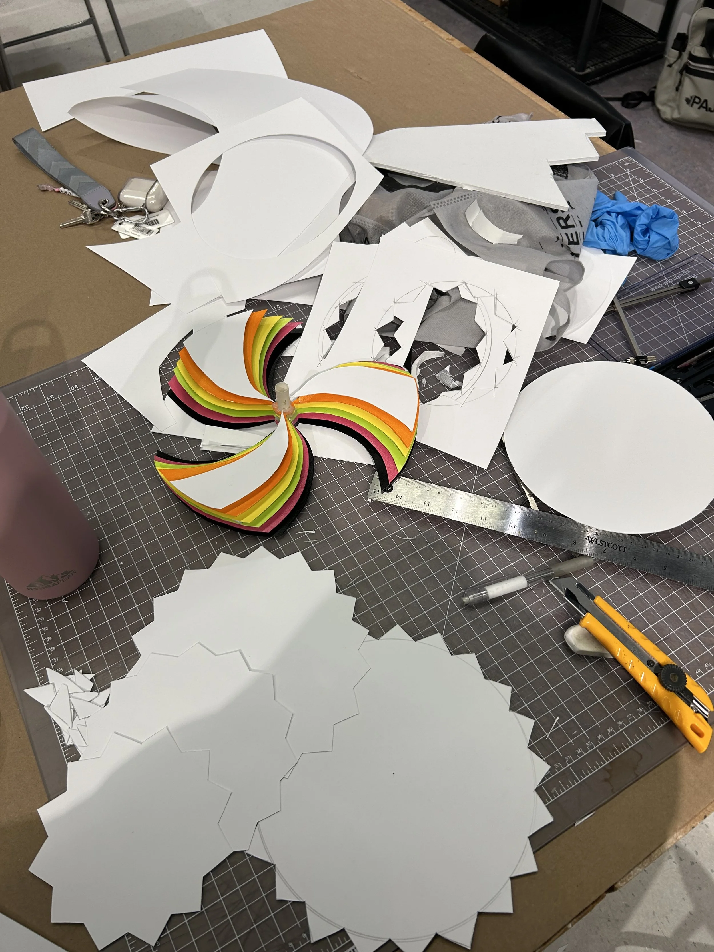 A cluttered workspace table with various cutout paper shapes, construction rulers, a utility knife, and some crafting materials, indicating an arts and crafts project in progress.