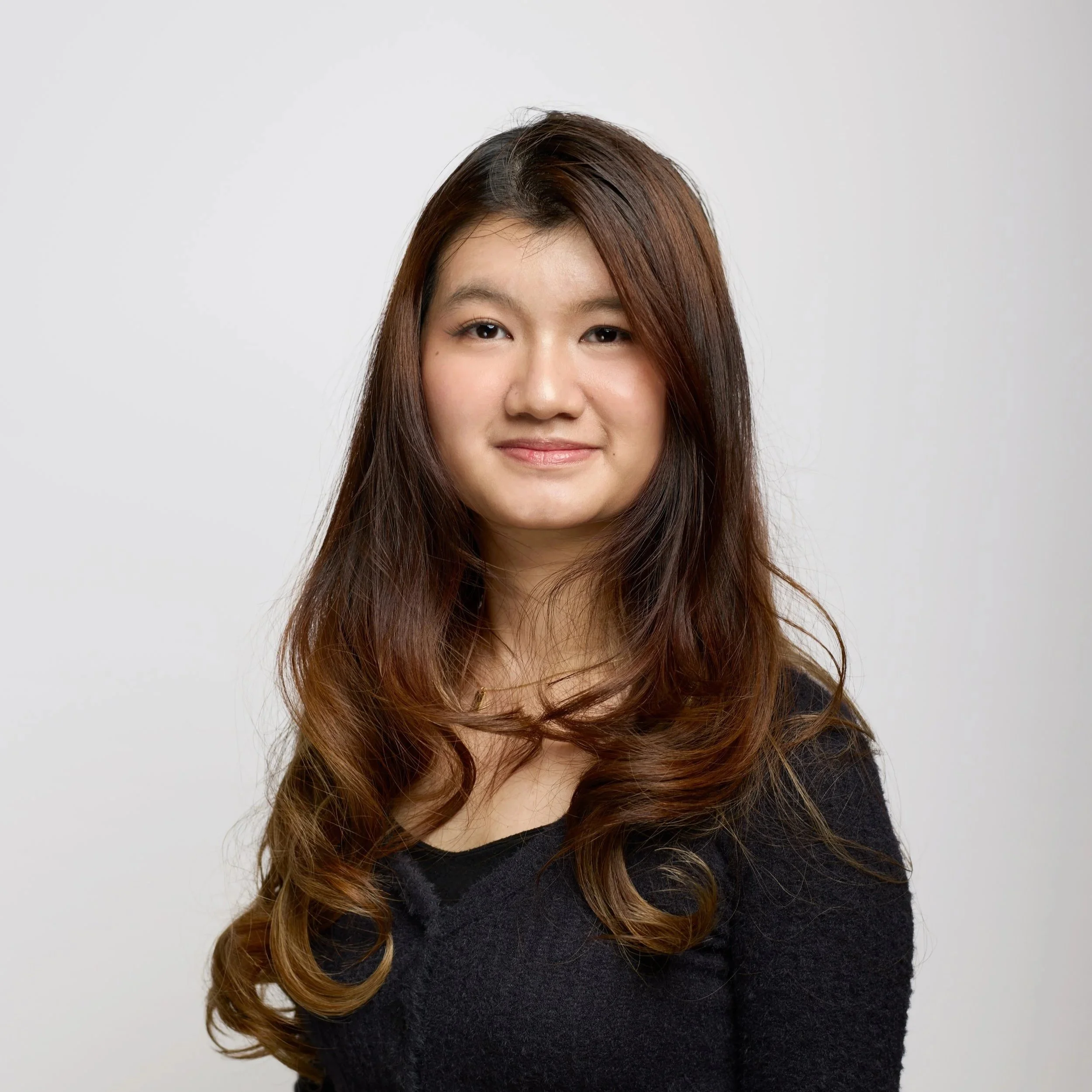 A young woman with long brown hair smiling at the camera, wearing a white shirt and a cream-colored jacket, against a black background.