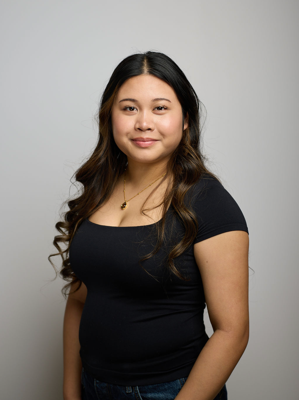 A young woman with long, wavy dark hair, smiling, wearing a black top and a gold necklace, standing against a plain light gray background.
