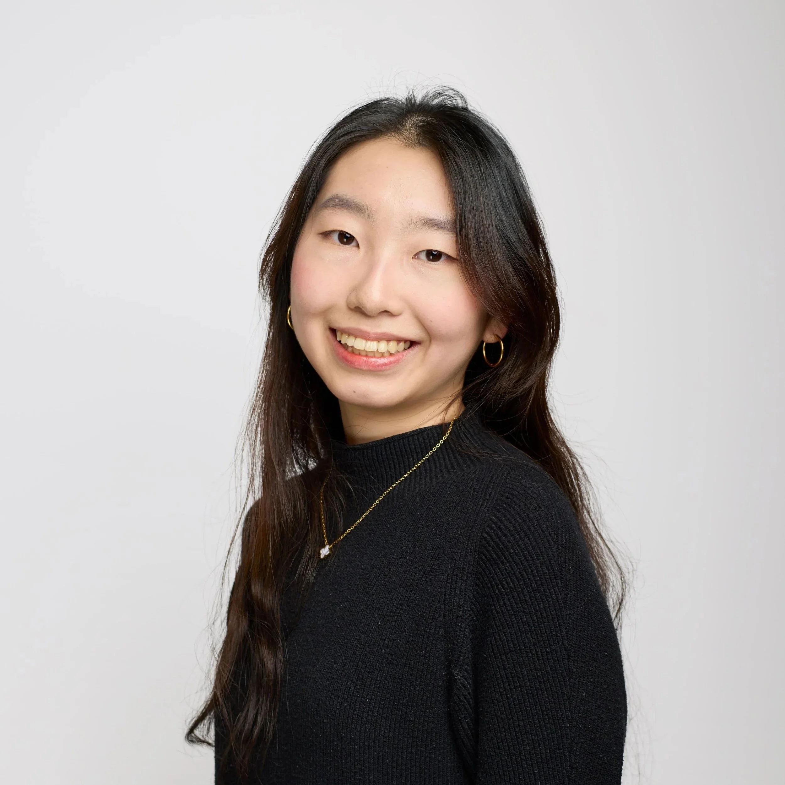 Close-up of a young woman with long dark hair, wearing hoop earrings and a black top, smiling against a plain light gray background.