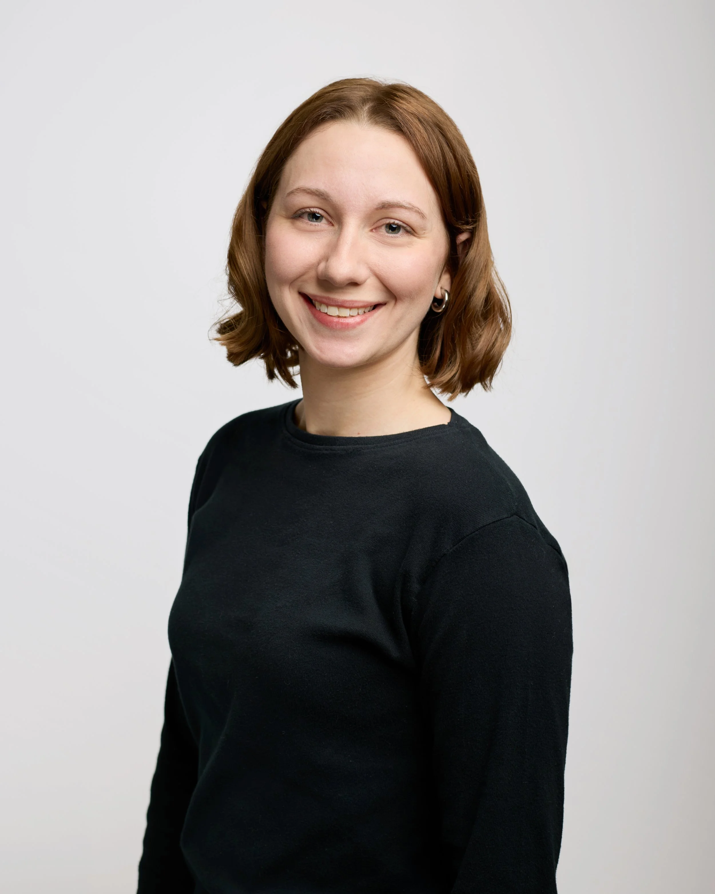 A young woman with shoulder-length reddish-brown hair, smiling, wearing a black top, standing against a plain white background.