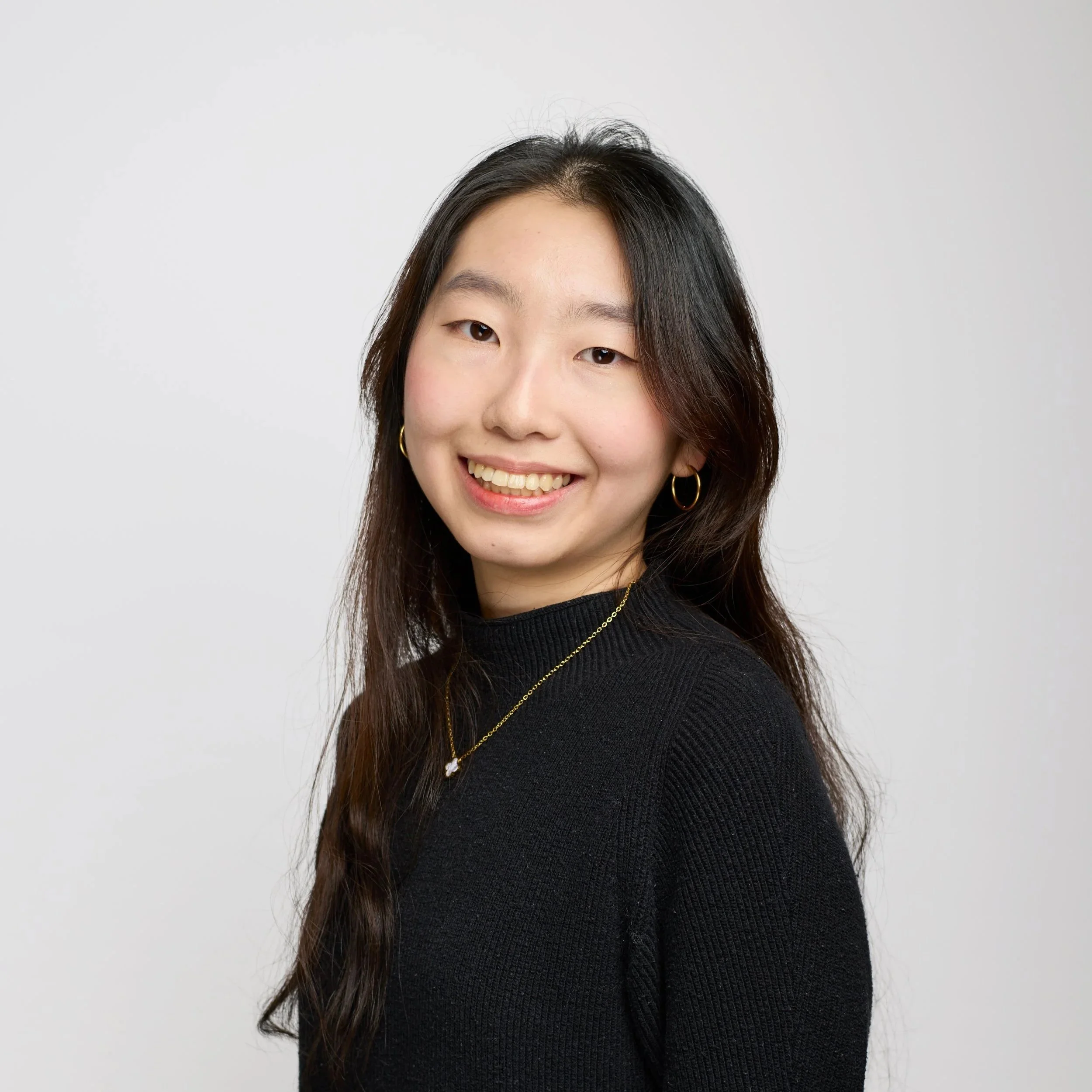 Young man with dark hair wearing a black sweatshirt and a silver necklace, posing against a black background.