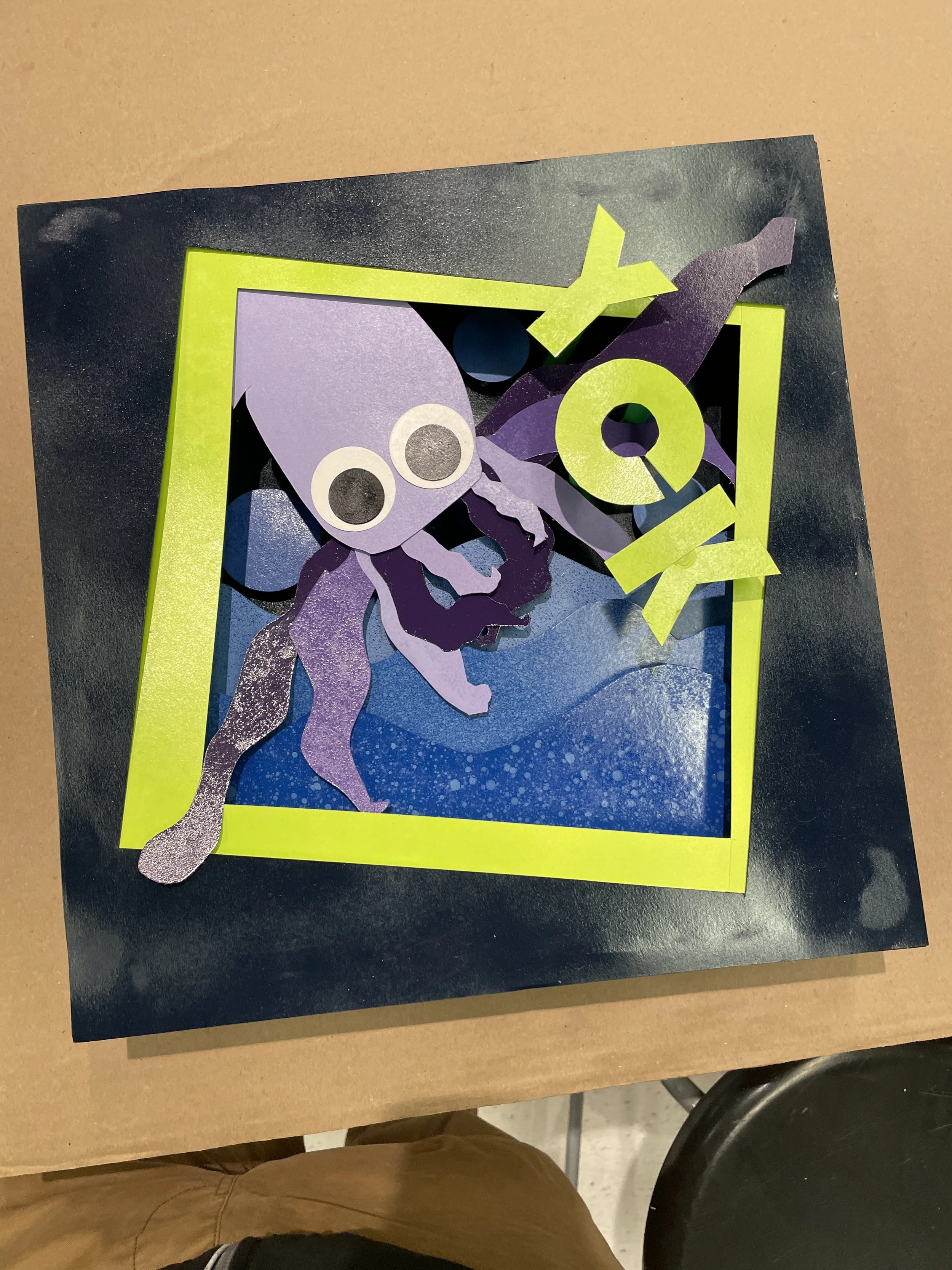 A 3D paper craft of a purple squid with big eyes and long tentacles inside a black frame with a yellow border, set against a dark background with blue accents.