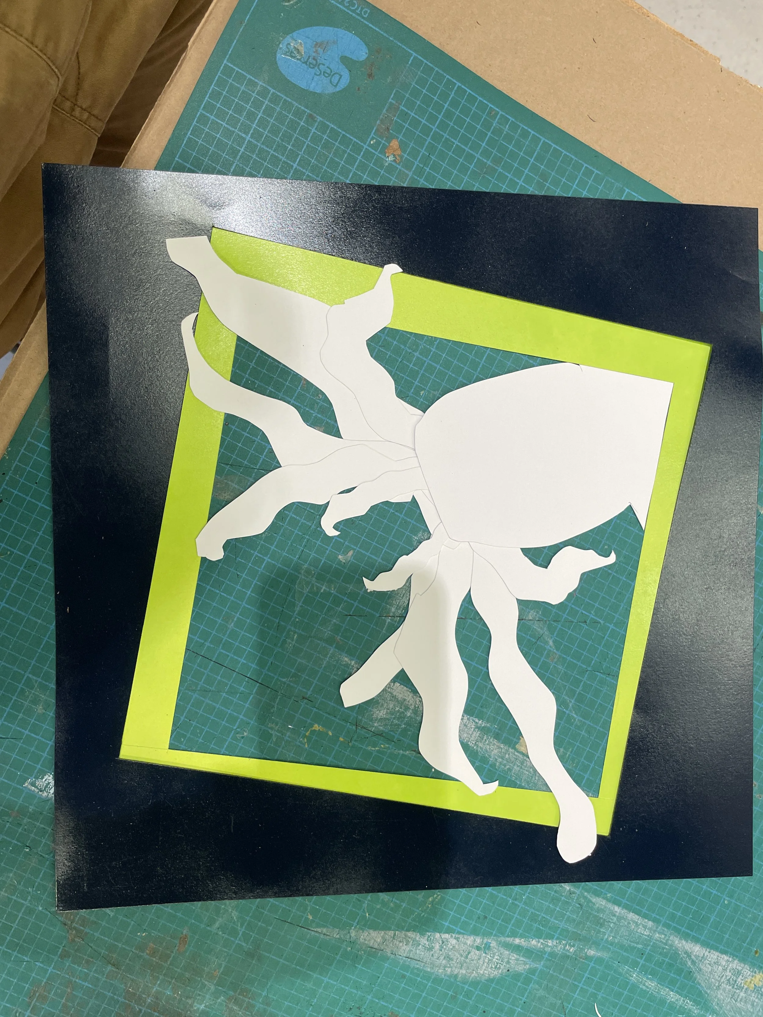 Paper craft of a white and green butterfly with black edges, attached on a black and green background, on a green cutting mat.
