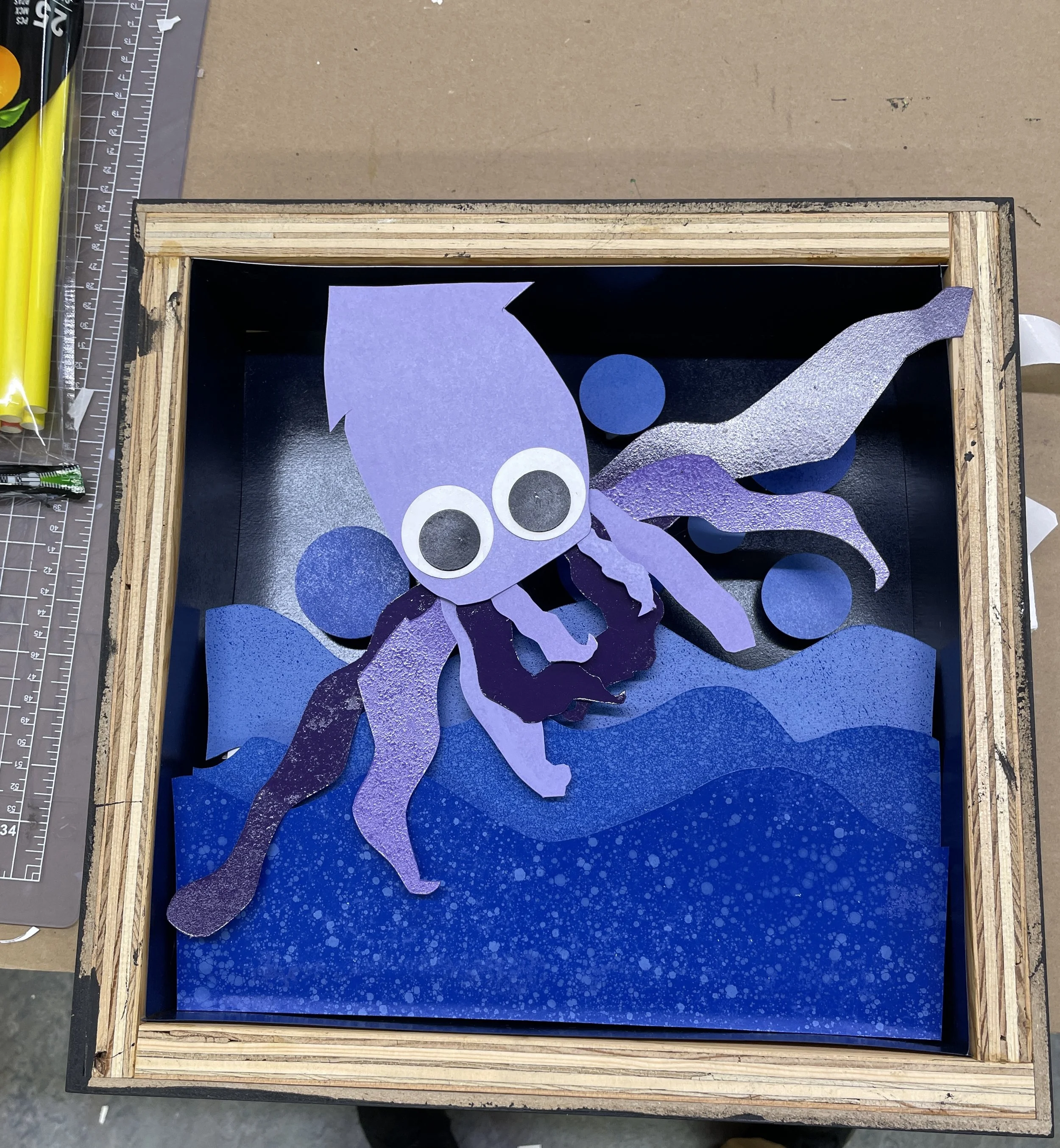 Colorful paper octopus craft with large googly eyes, placed inside a black box frame on a workspace with measurement tools on the side.