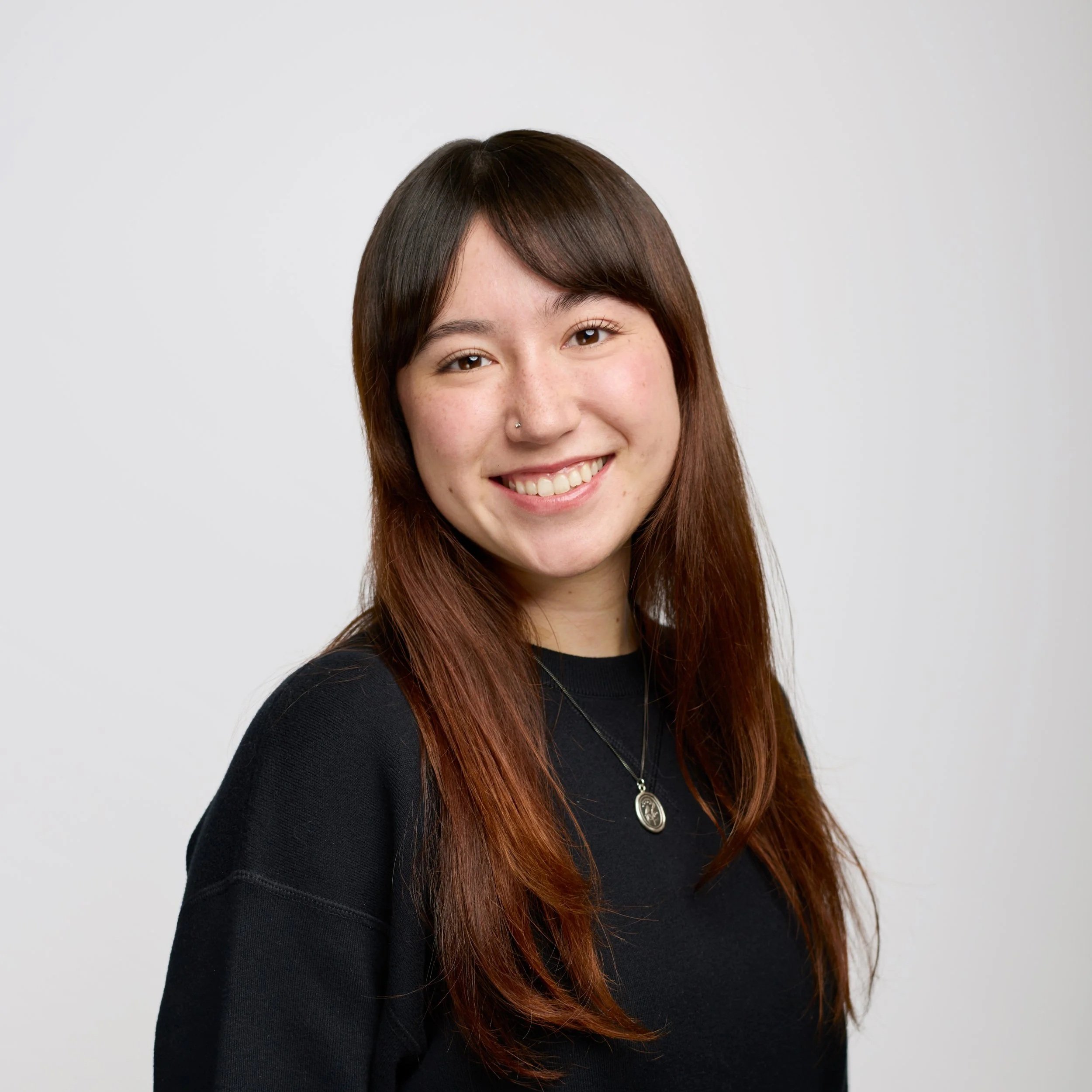 A young woman with shoulder-length dark brown hair, smiling, wearing a cream-colored sweater and a pendant necklace, against a black background.