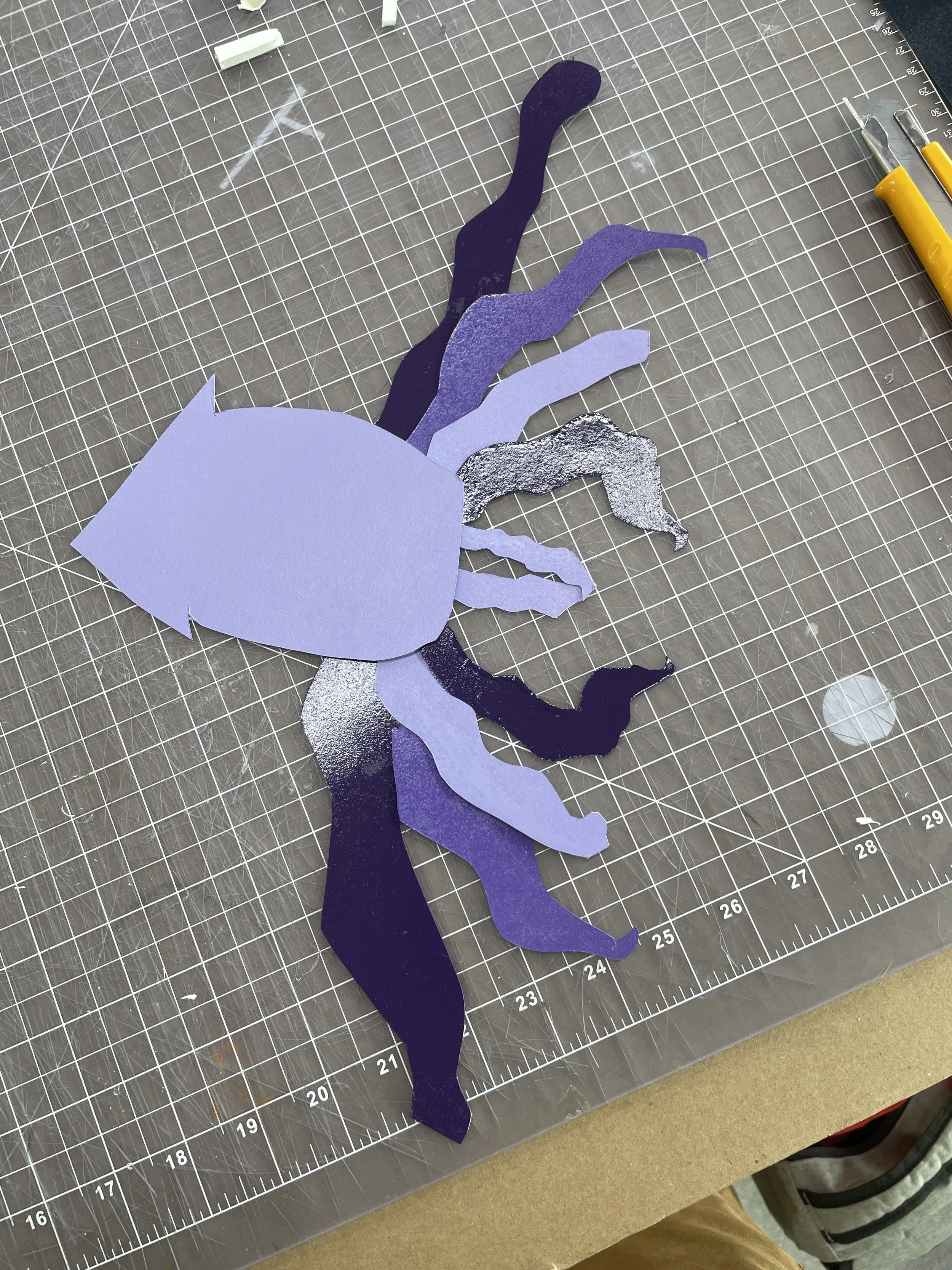 Several layers of purple and metallic paper cutouts arranged on a grid cutting mat. A yellow utility knife and a precision craft knife are visible on the right side of the mat.