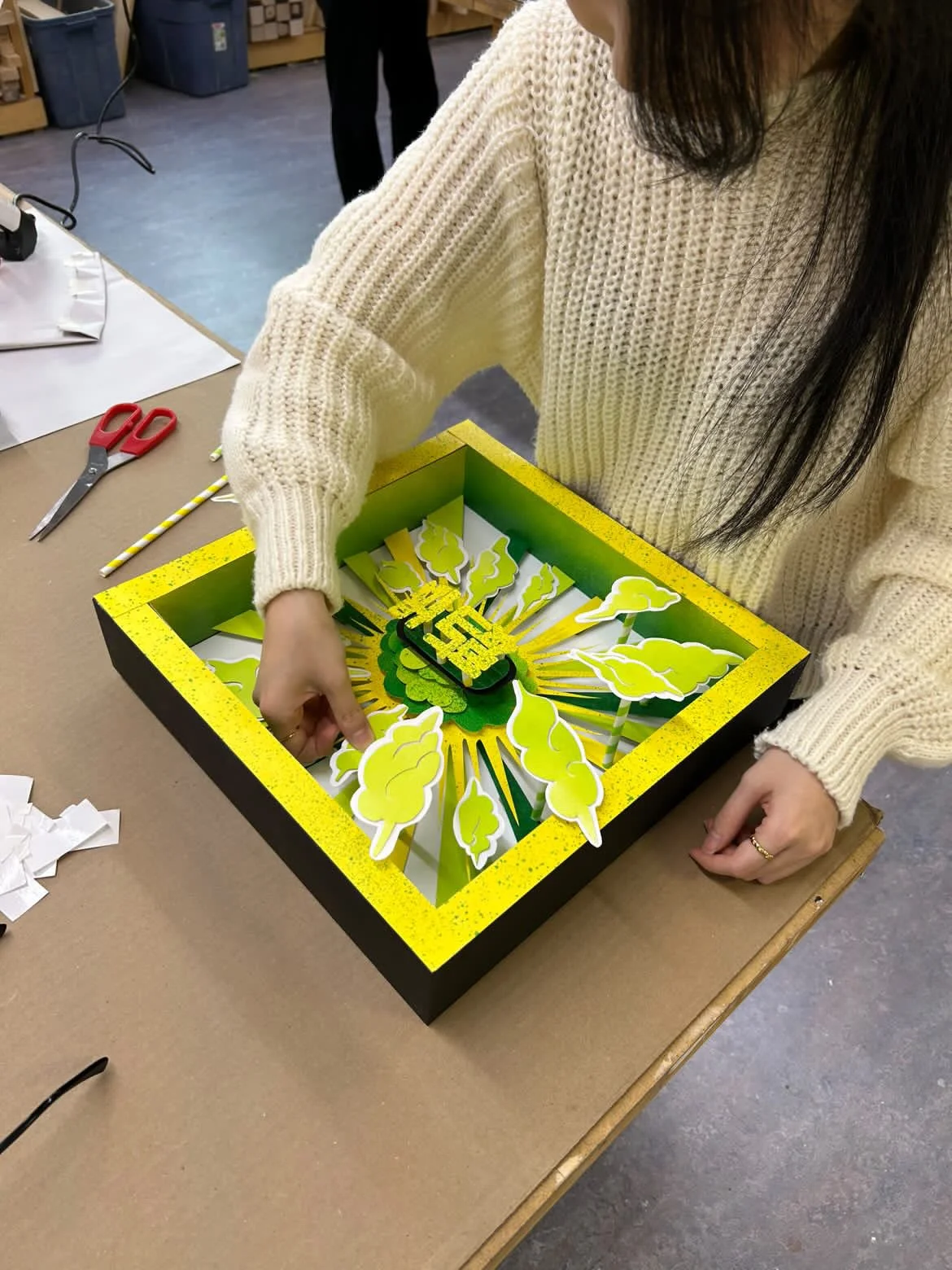 Person assembling a decorative paper clock with green, yellow, and black colors, featuring abstract leaf patterns and 3D elements, on a craft table.