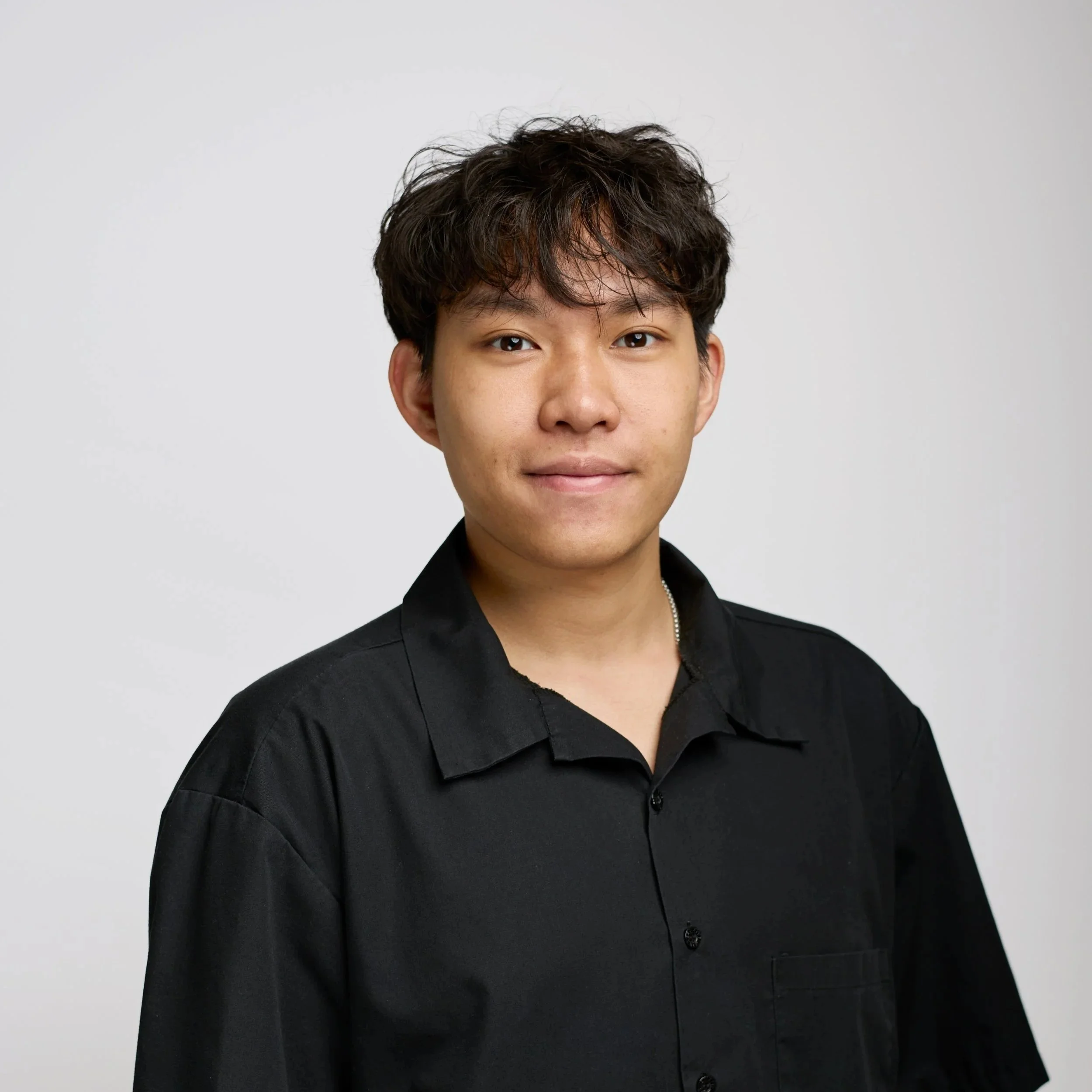 Portrait of a young man with short black hair, wearing a black sweatshirt and silver chain necklace, smiling against a black background.
