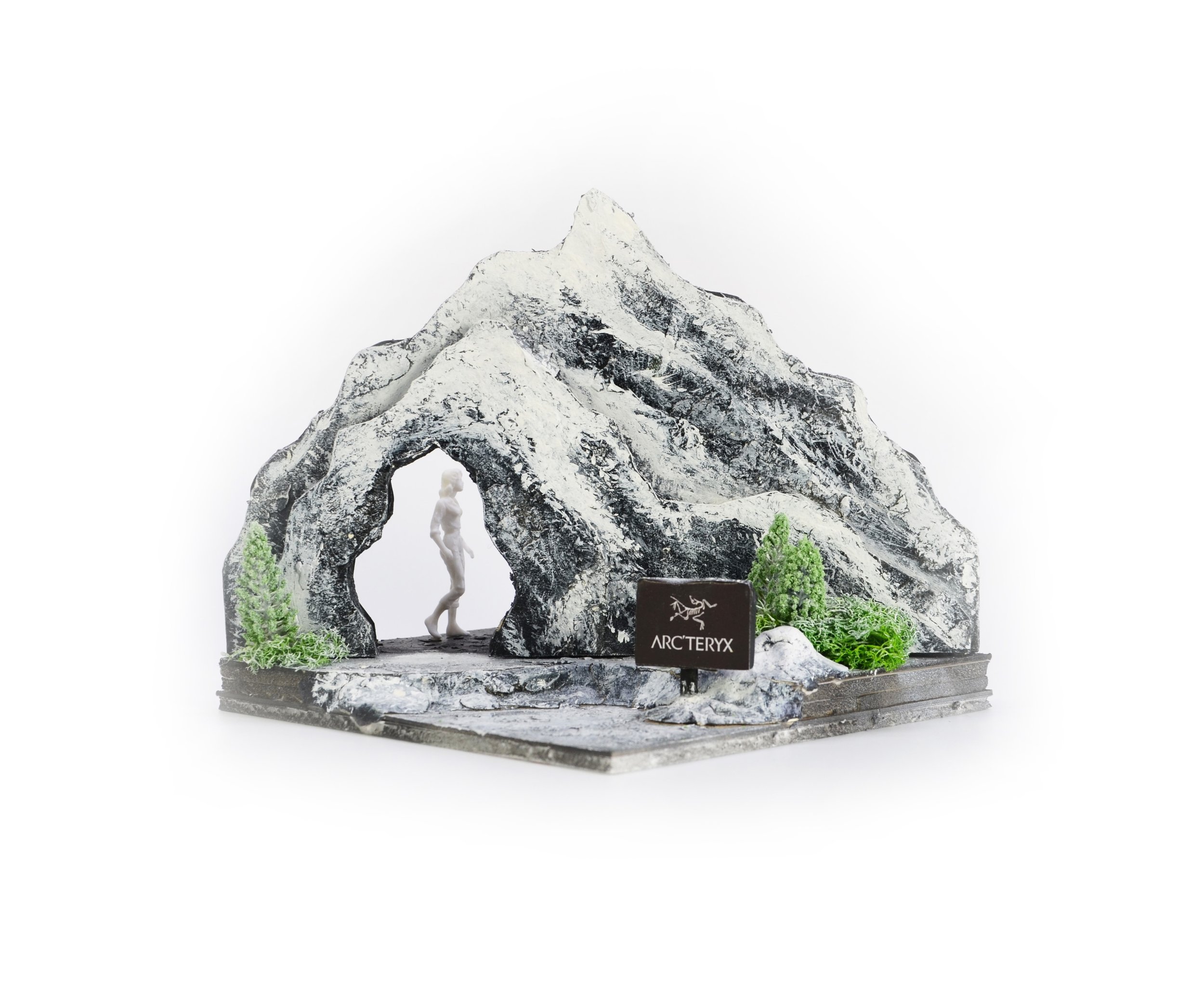 A decorative miniature sculpture of a mountain with an arch underneath, featuring a small figurine of a person standing at the base, surrounded by green moss and labeled Arc'teryx.