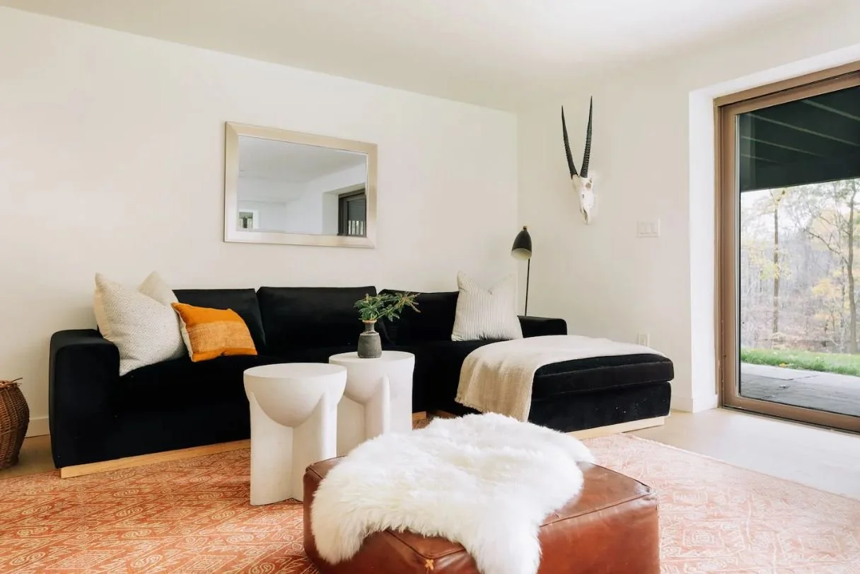 Living room with a black sectional sofa decorated with white and orange pillows, a white fluffy throw blanket, a mirror on the wall, a wall-mounted animal skull with antlers, a floor lamp, and a sliding glass door showing an outdoor view of trees.