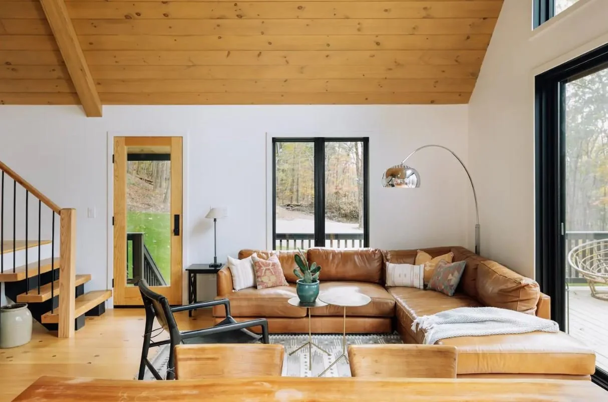 Living room with a brown leather sectional sofa, a black armchair, a wooden dining table, and wood ceiling. Large windows and sliding glass door show a wooded outdoor area.
