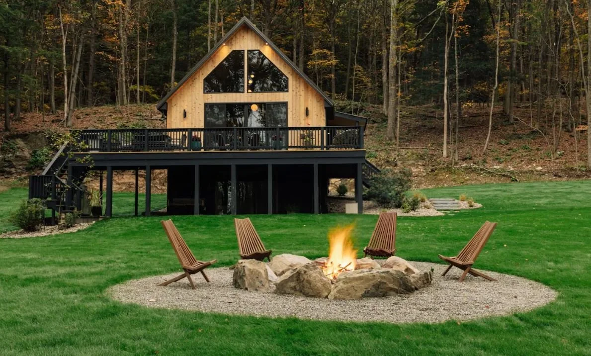 A backyard with a fire pit surrounded by four wooden chairs, a large green lawn, and a modern A-frame house with a black deck, set against a wooded hillside.