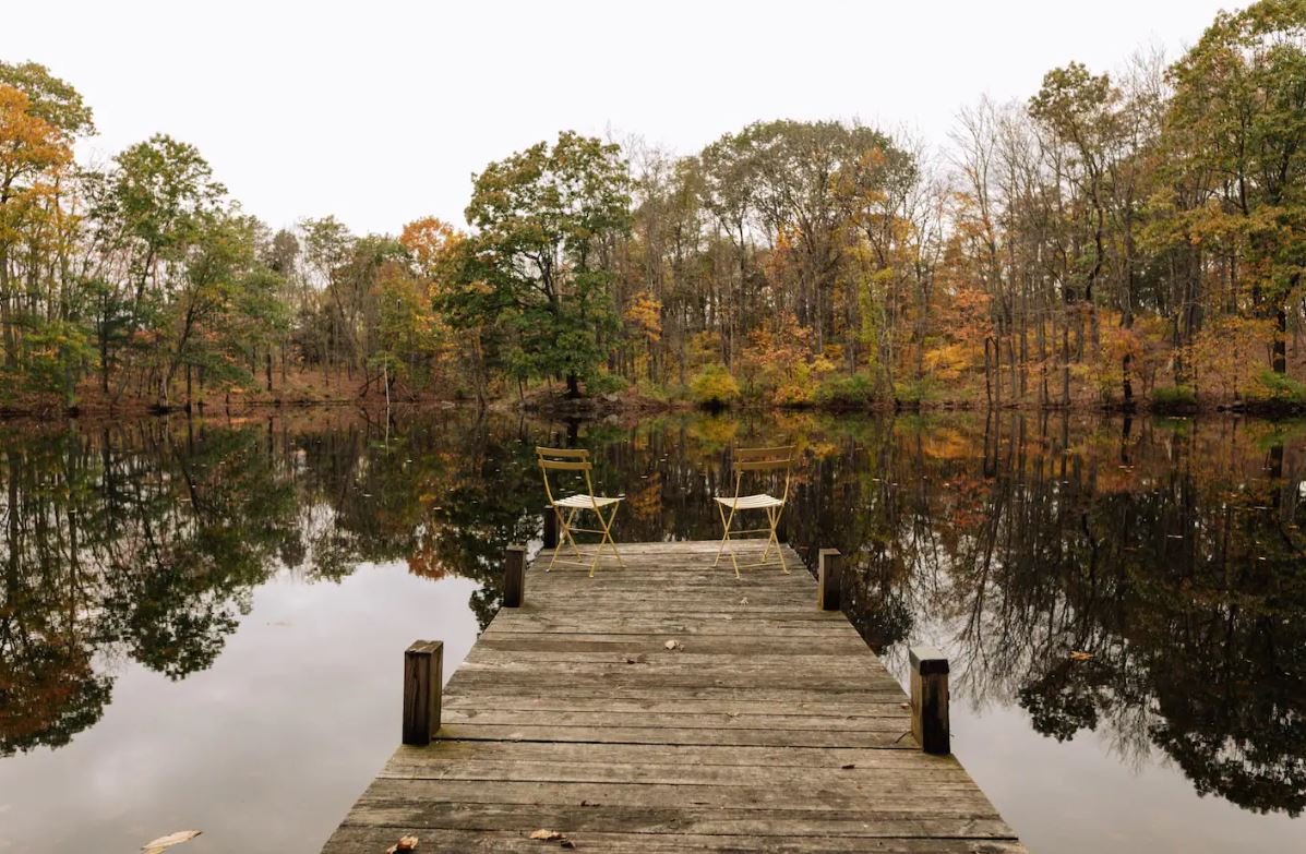 Two chairs on a wooden dock overlooking a calm lake surrounded by autumn-colored trees.