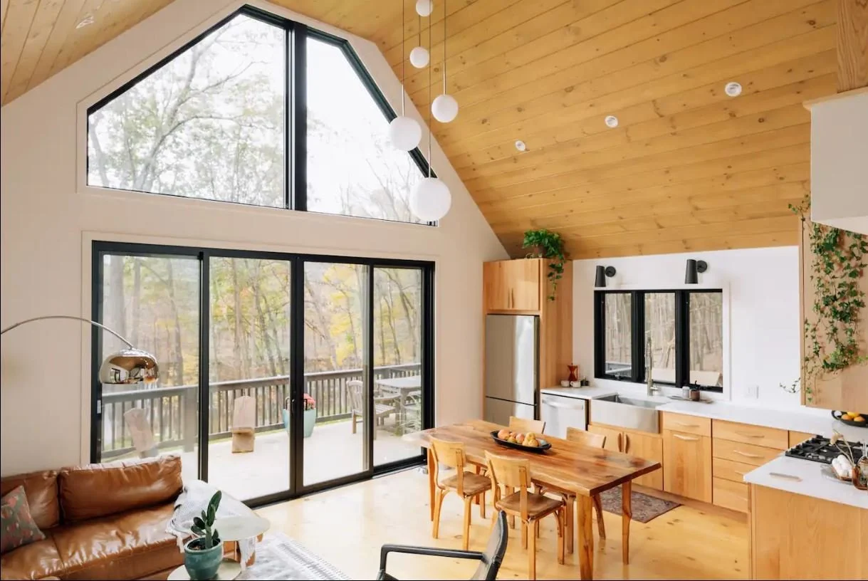 A modern kitchen and living area with large windows and sliding glass doors leading to a balcony, featuring wooden ceilings and furniture, a leather sofa, and decorative plants.
