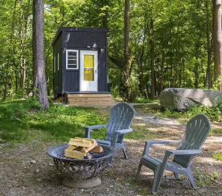 A small black cabin with white trim and a yellow door in a wooded area. In front, there are two blue Adirondack chairs and a metal fire pit with firewood inside, set on a dirt clearing surrounded by trees.