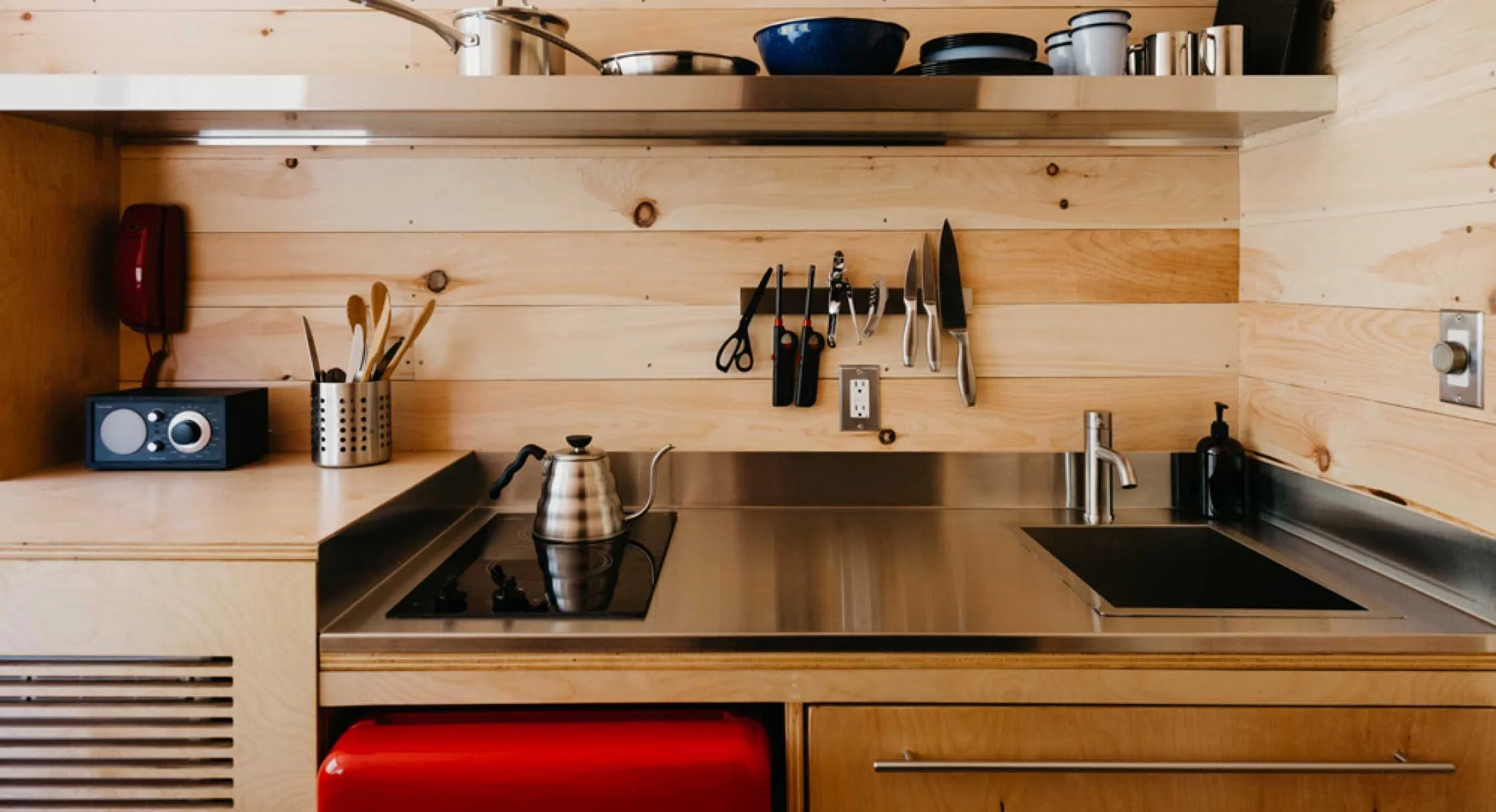 A modern kitchen with light wood walls and cabinets, featuring a stainless steel sink, a stovetop with a kettle, and a wall-mounted knife and scissors rack. There is a small speaker, a utensil holder, and a red appliance below the counter.