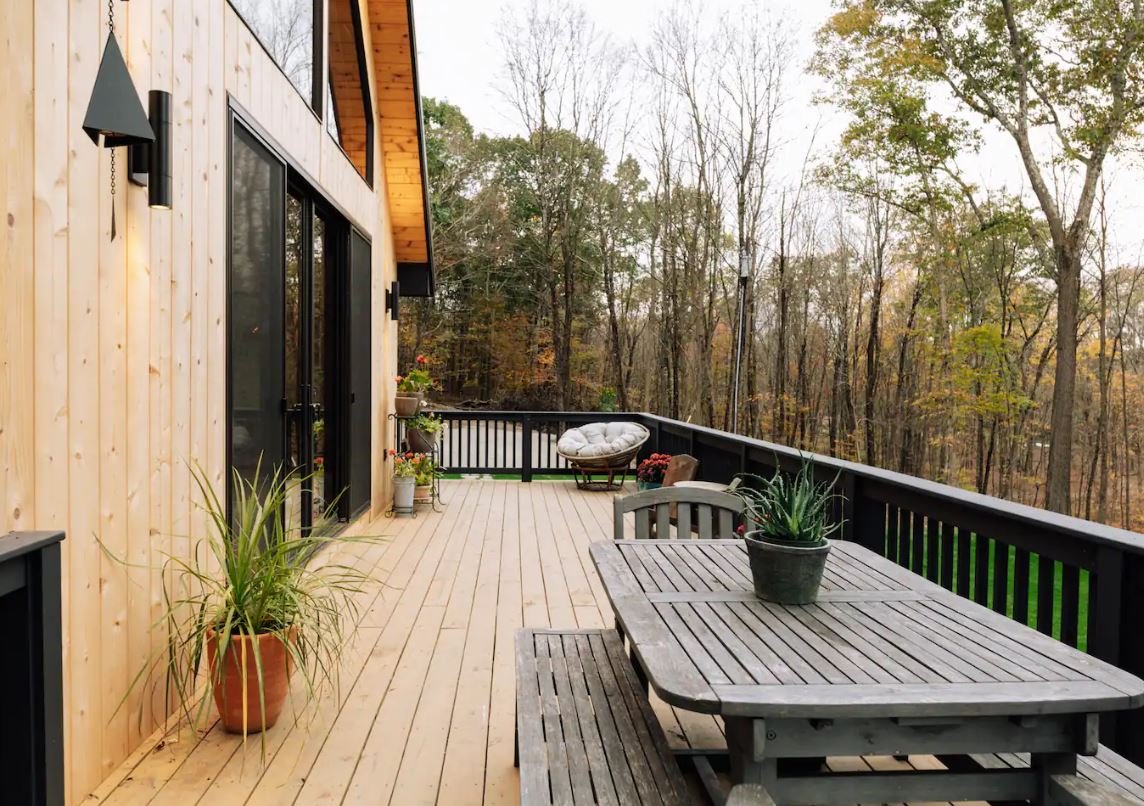 Wooden deck attached to a house with outdoor furniture, potted plants, and a forested background.