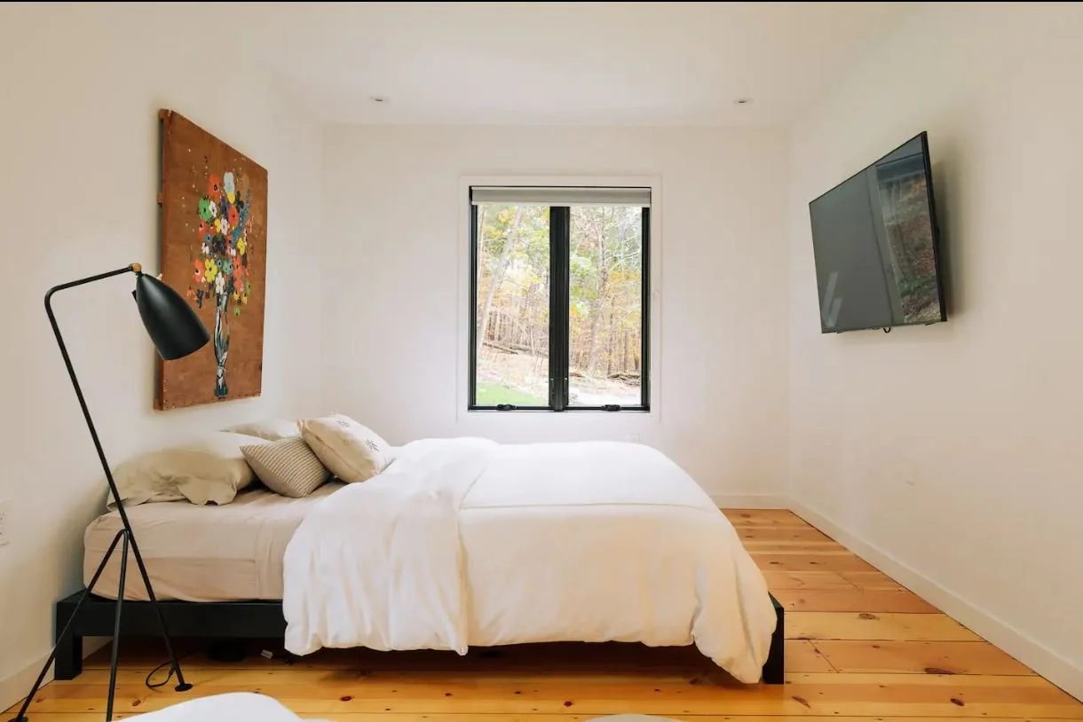 A minimalist bedroom with a bed covered in white linens, a black floor lamp on the left, a colorful floral artwork above the head of the bed, a window with a view of trees outside, and a flat-screen TV mounted on the right wall.