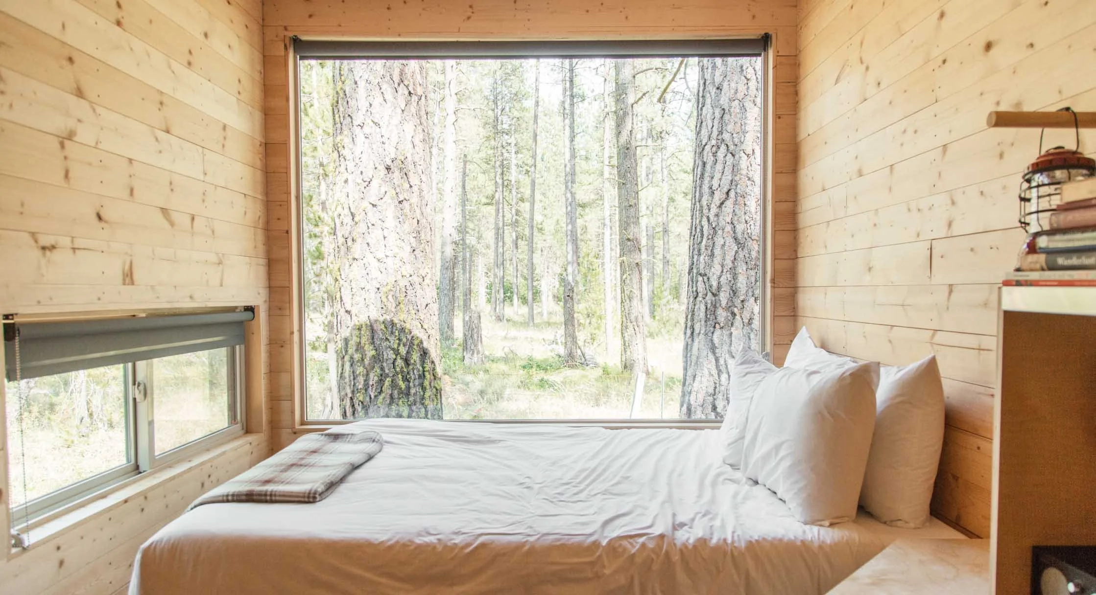 A cozy bedroom with wooden walls and large windows showing a forest outside, featuring a bed with white bedding and pillows, and a small bookshelf with books on the right side.