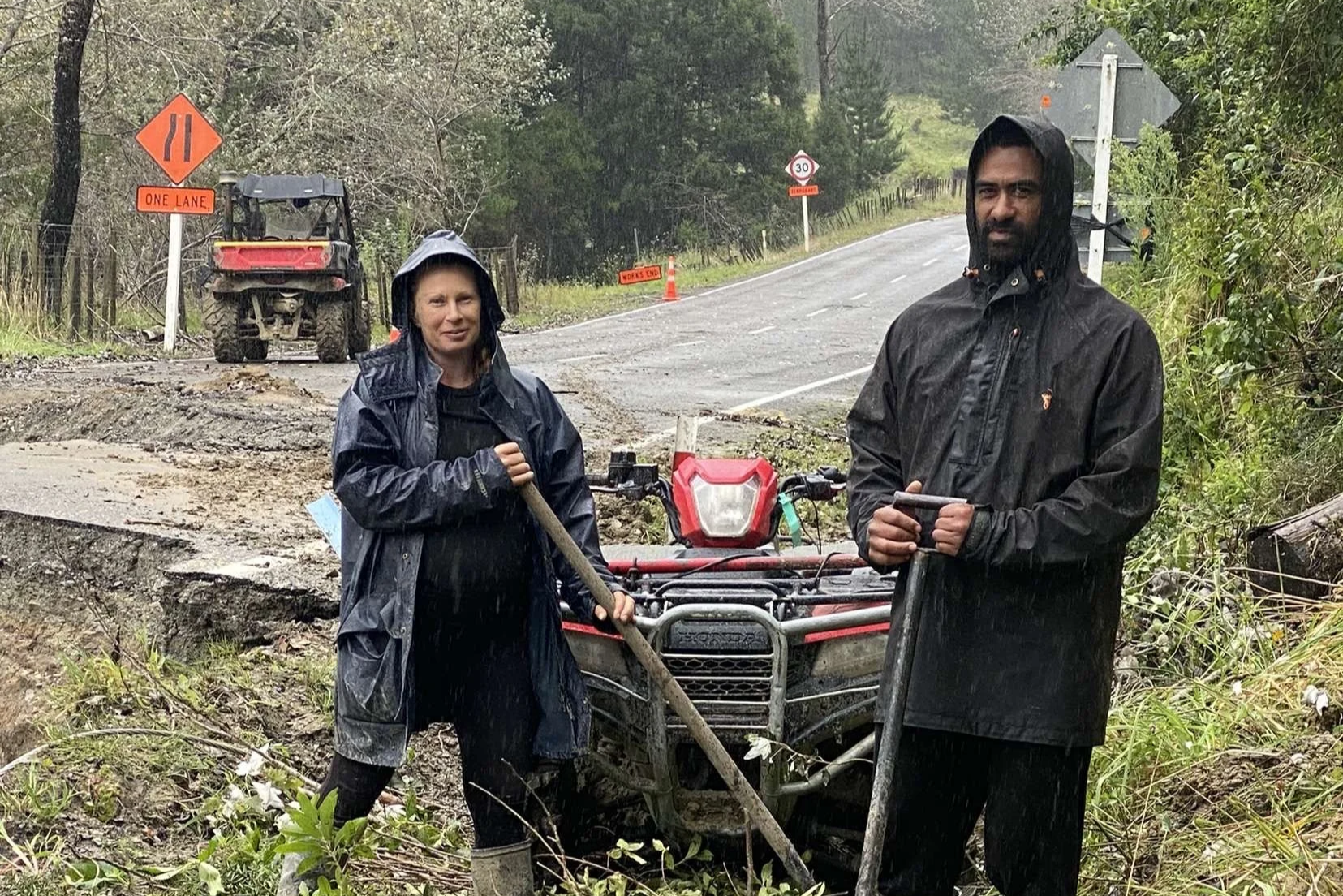 Two people in rain gear fixing a muddy road, with ATVs and a sign indicating a blocked road.