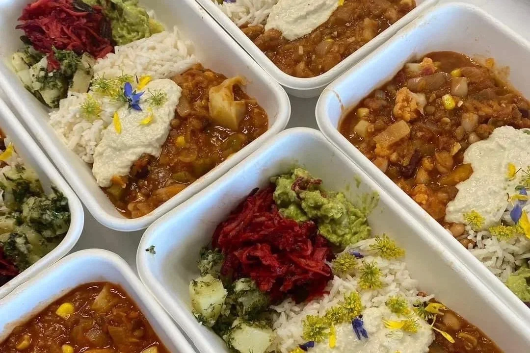 Multiple white meal containers filled with rice, lentil stew, guacamole, shredded beets, and diced onions, garnished with edible flowers and herbs.