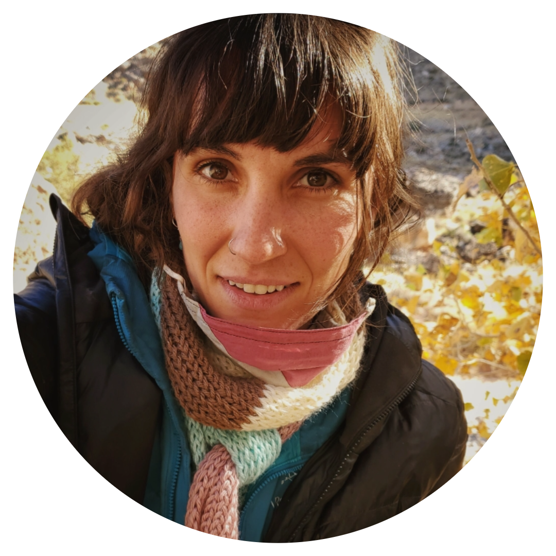 Close-up of a woman outdoors, smiling, with brown hair, wearing a colorful scarf, jacket, and a face mask pulled down, surrounded by fall foliage.